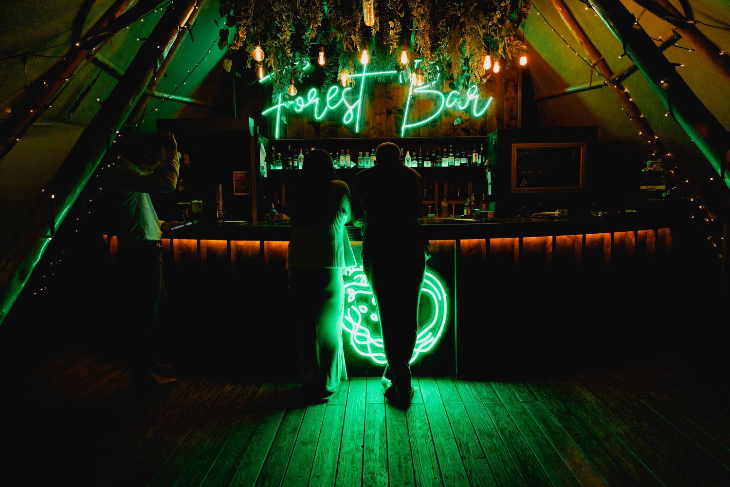 Guest stands before glowing green Forest Bar neon sign inside atmospheric tipi wedding venue.