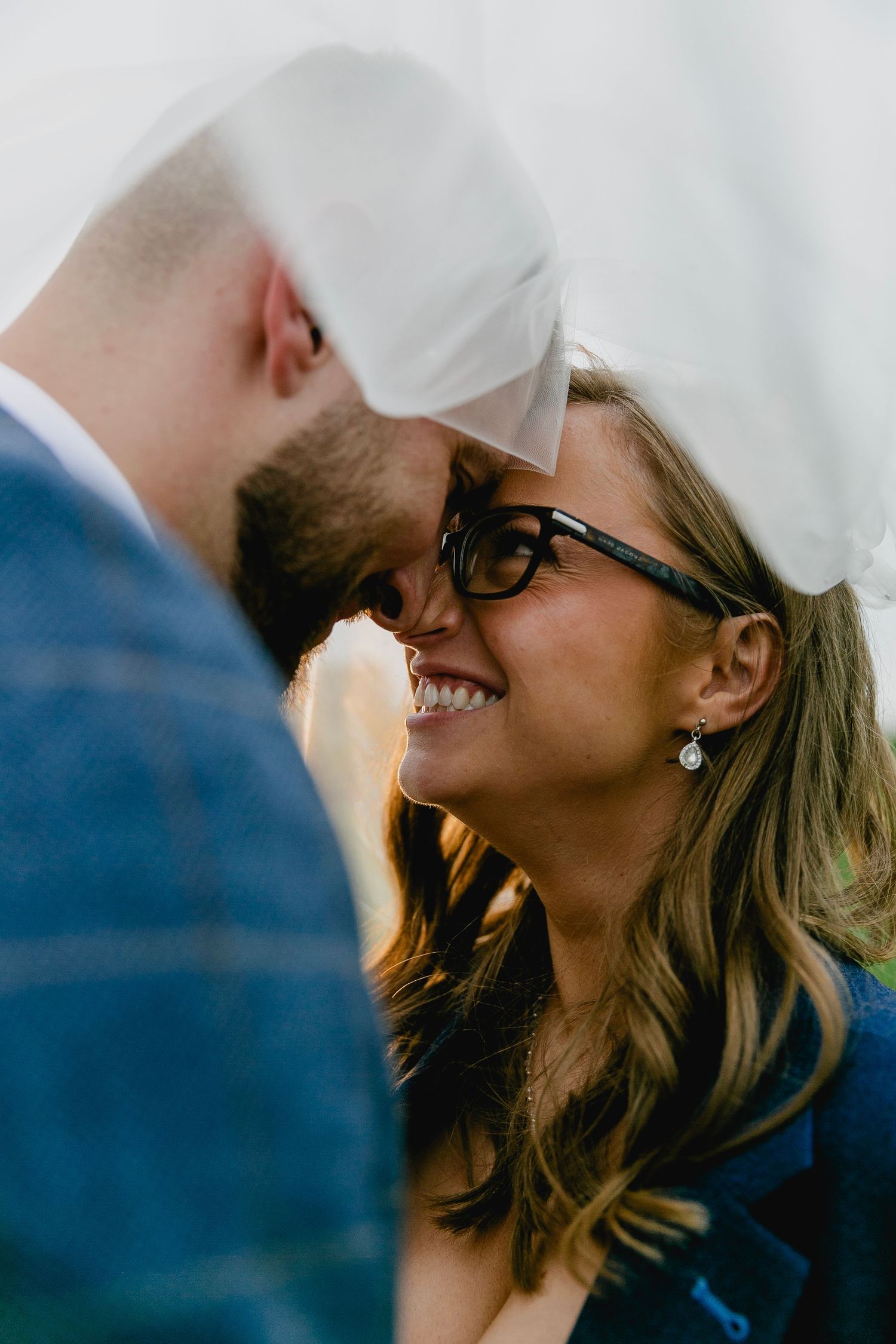 Bride wearing glasses laughing joyfully with groom under wedding veil