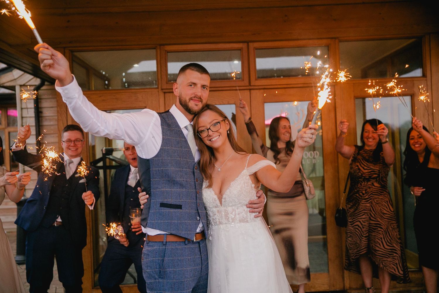 Newlywed couple smiling and holding sparklers surrounded by guests at their wedding celebration.