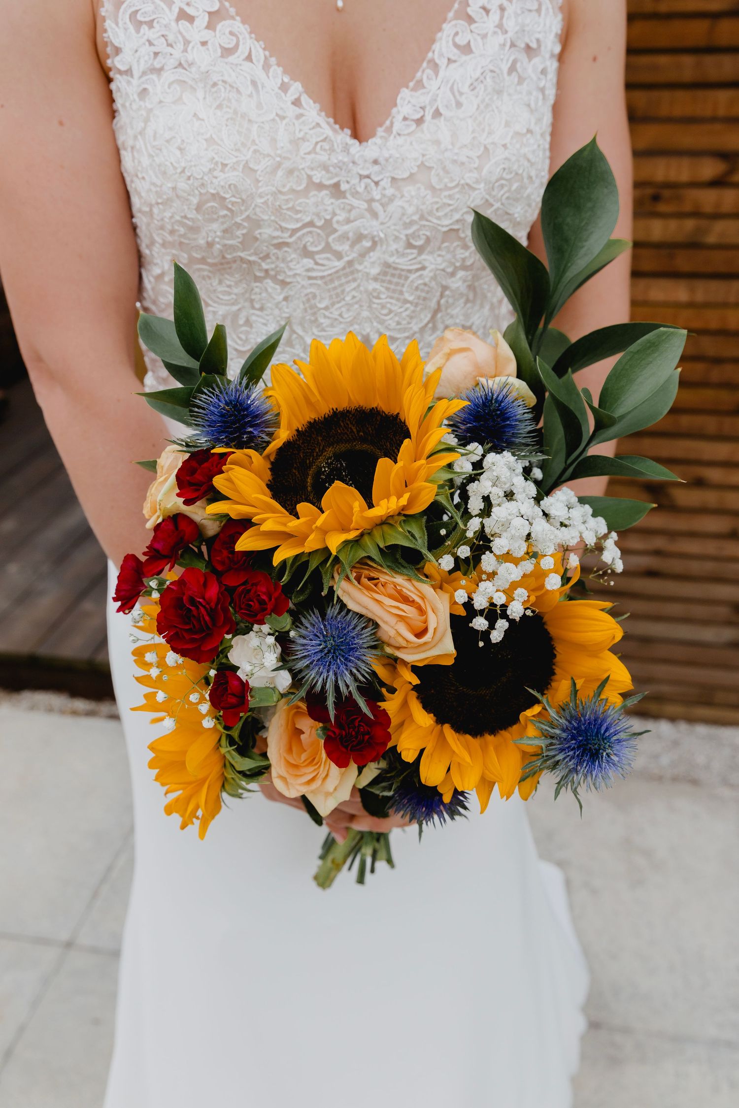 Bride holding vibrant sunflower wedding bouquet with red roses, blue thistles, and baby breath.