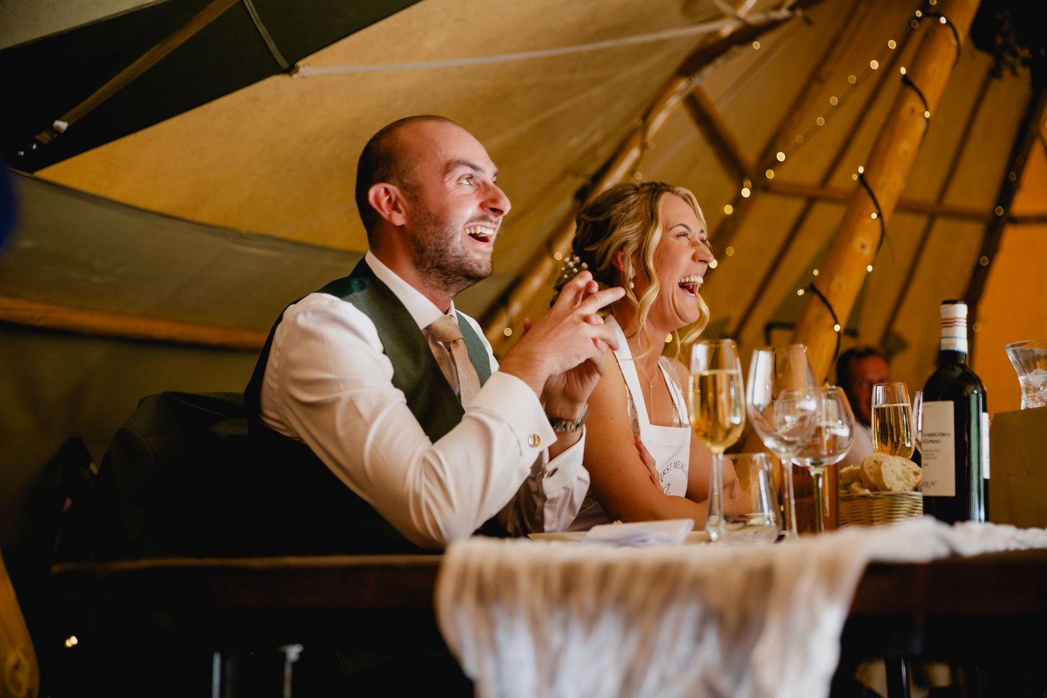 Laughing bride and groom enjoy speeches at wedding reception inside a fairy-lit tipi.