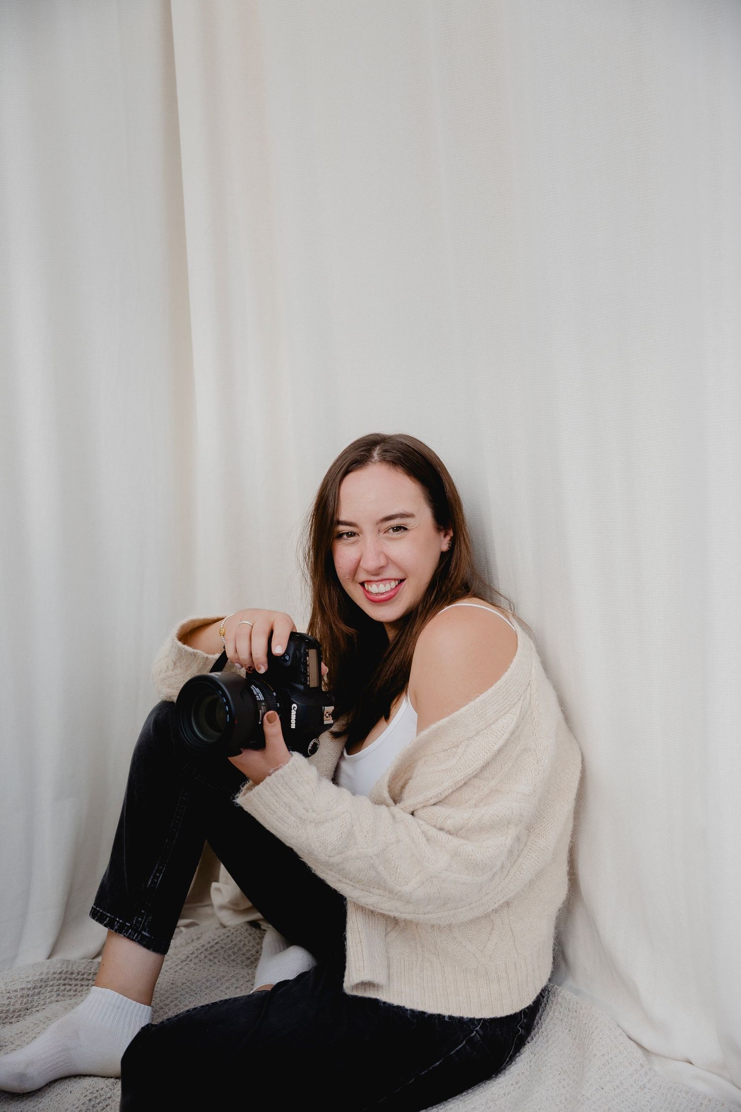 A smiling young woman sits against white curtains holding a DSLR camera in a beige cardigan.