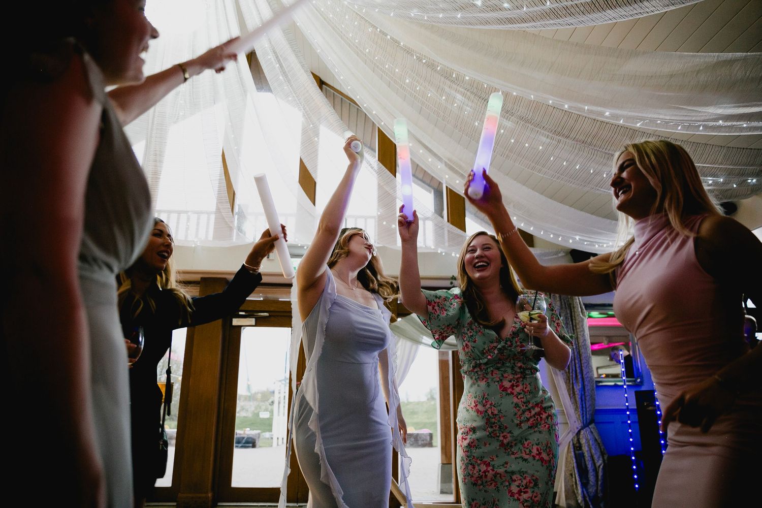Group of women celebrating at a wedding reception, holding glowing light sticks and dancing together.