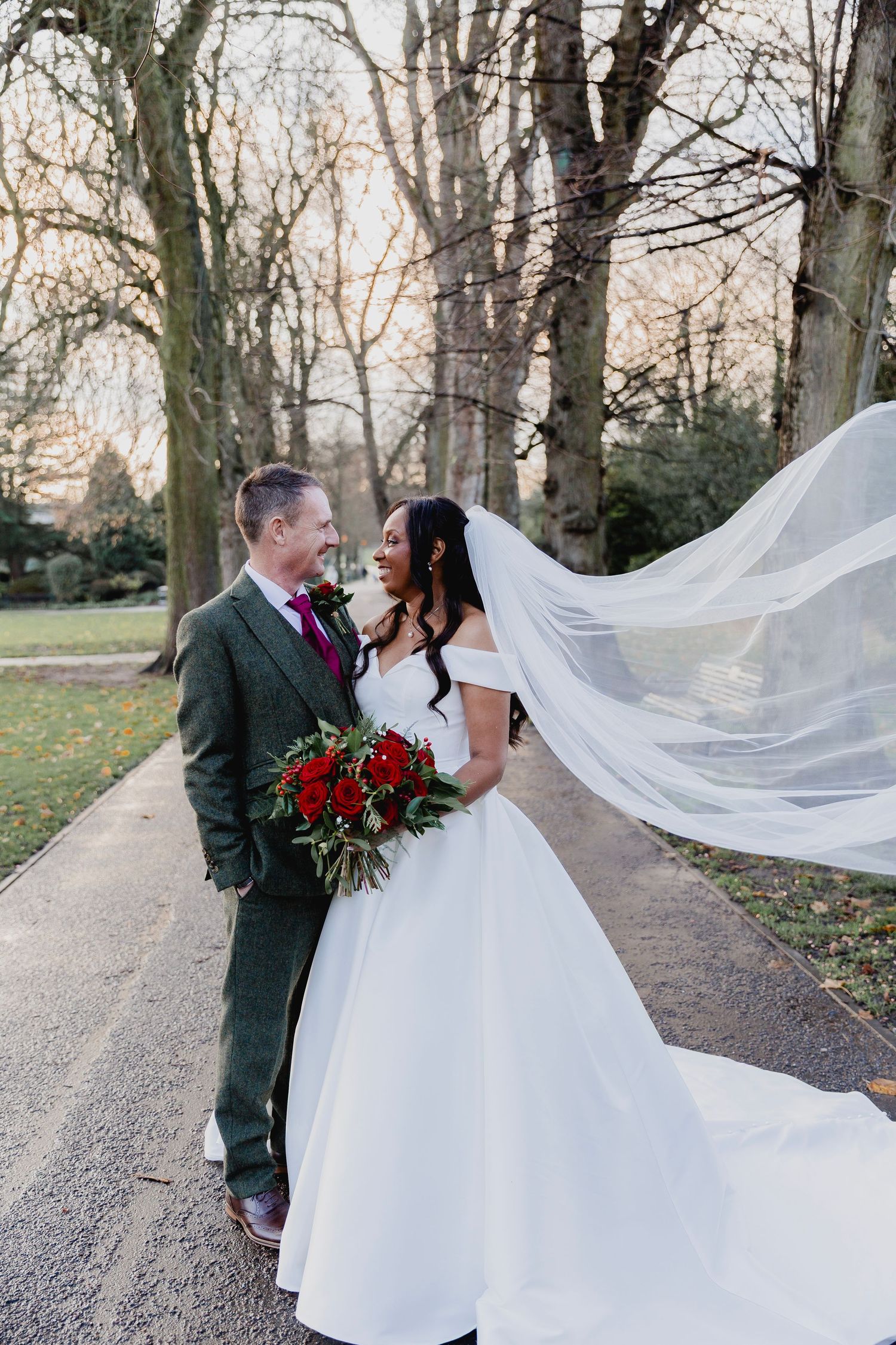 A bride and groom stand together outdoors as her long veil flows dramatically in the wind.