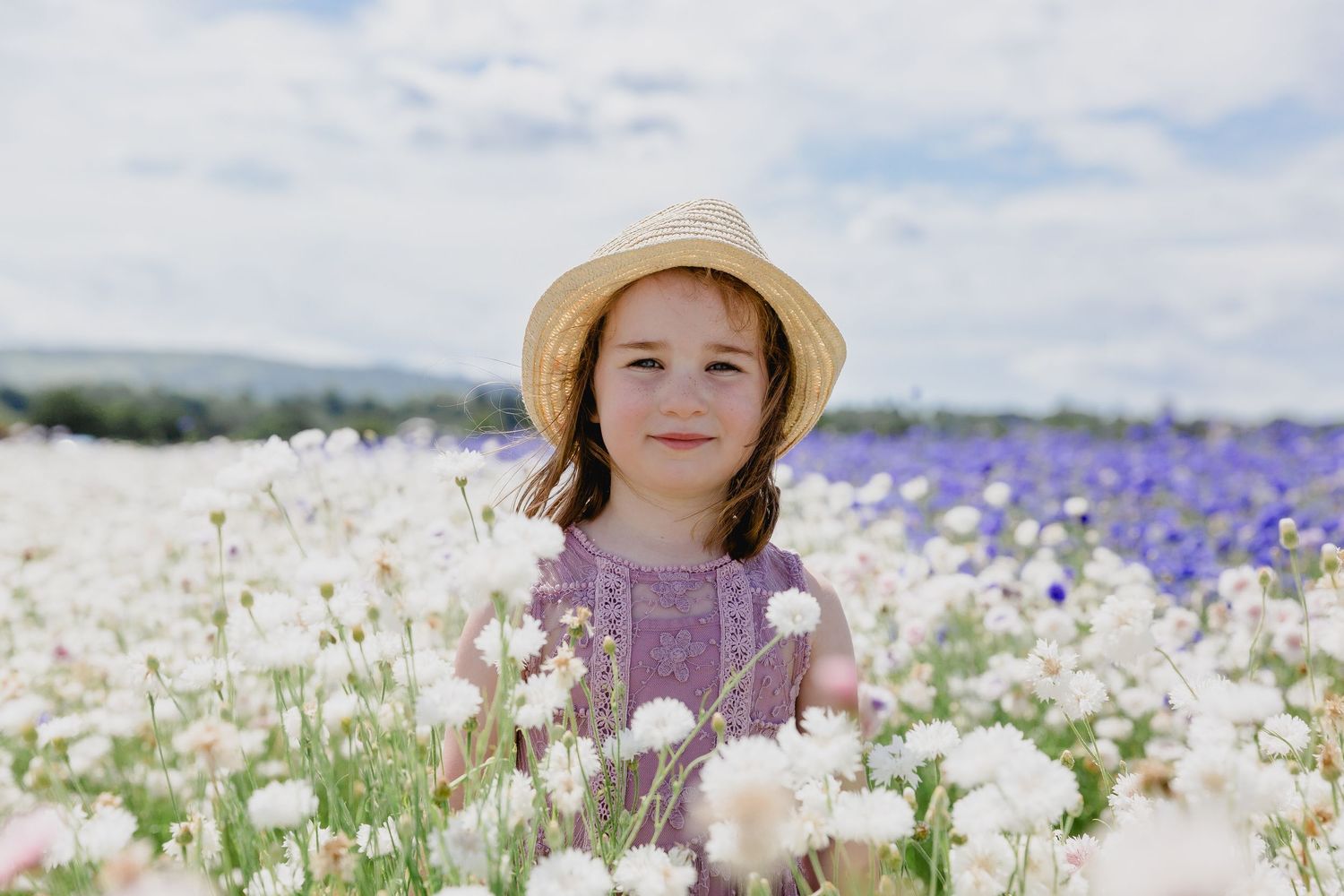A young girl in a straw hat smiles while standing in a vibrant field of white and purple flowers.