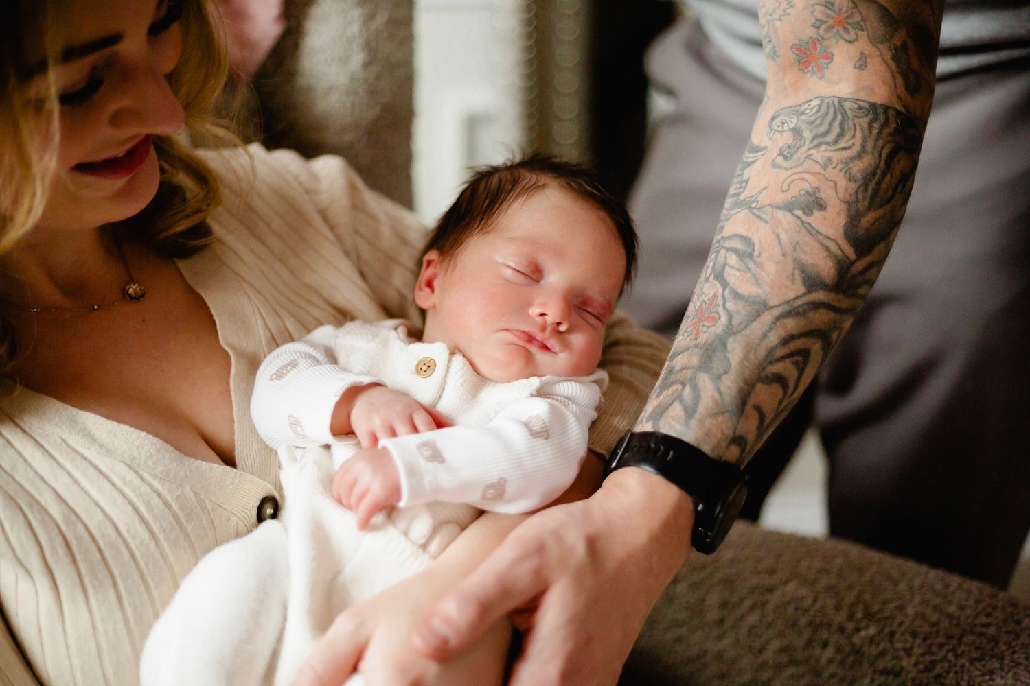 A sleeping newborn baby in white onesie cradled in arms, with a tattooed parent nearby.
