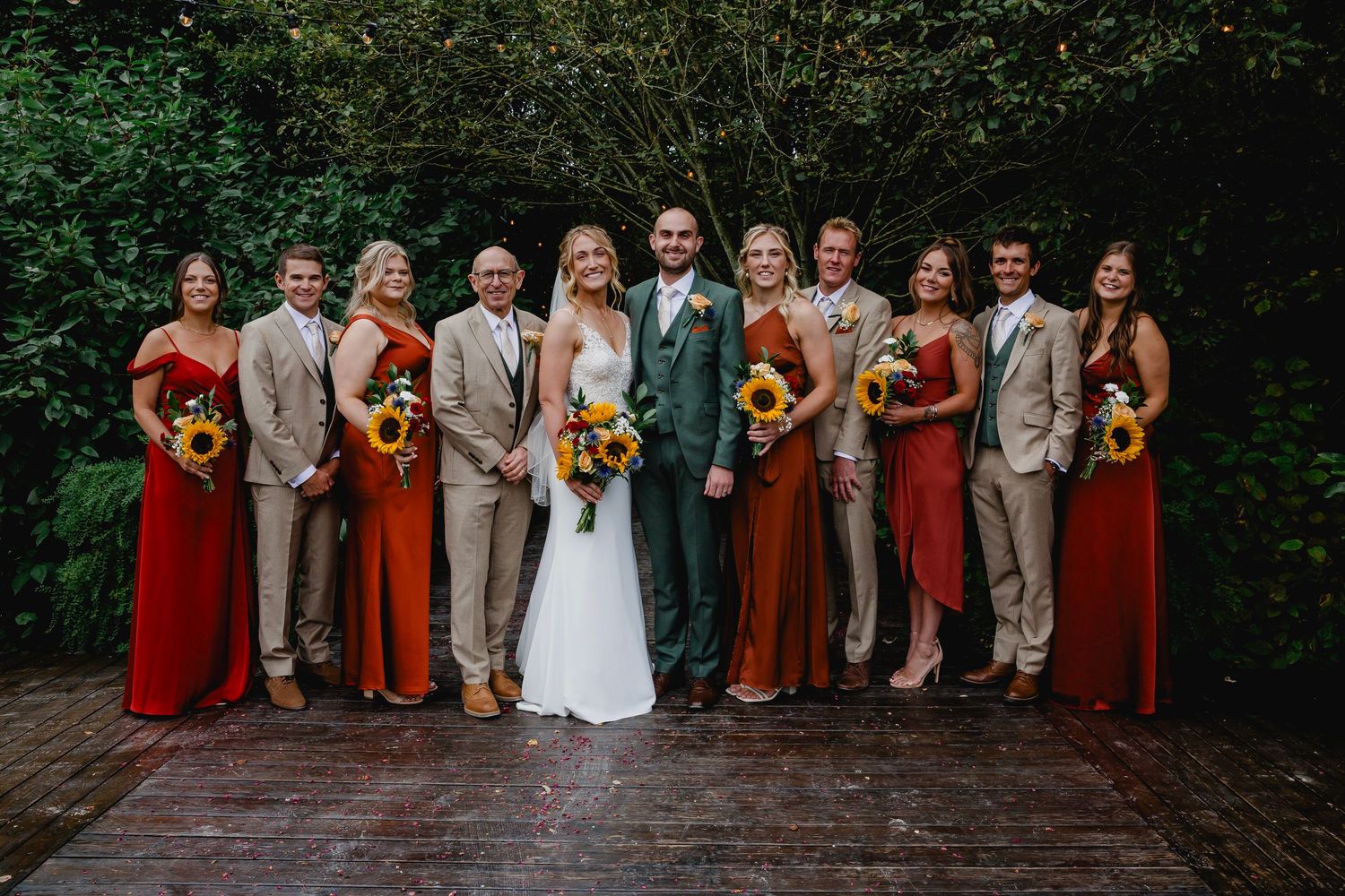 Wedding party in red and tan dresses with sunflower bouquets posing with bride and groom outdoors.