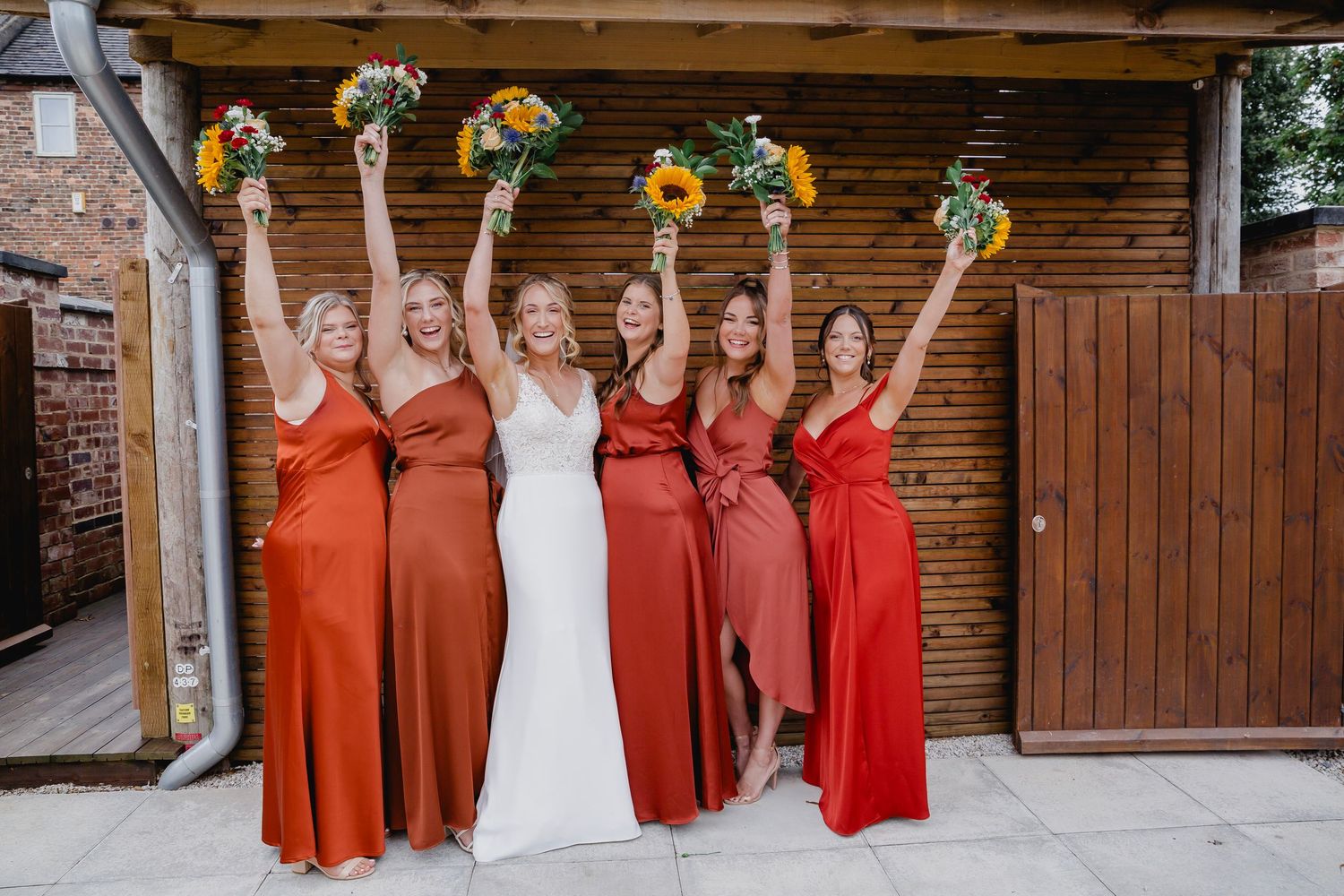 Bride and five bridesmaids in orange and red dresses raising sunflower bouquets outside rustic barn venue.