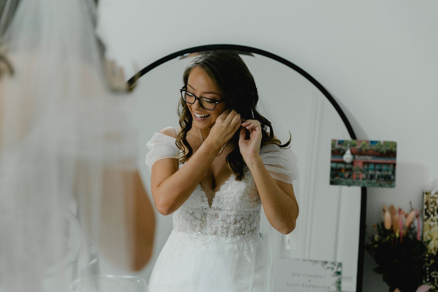Bride smiling while putting on earrings in front of round mirror during wedding preparations.