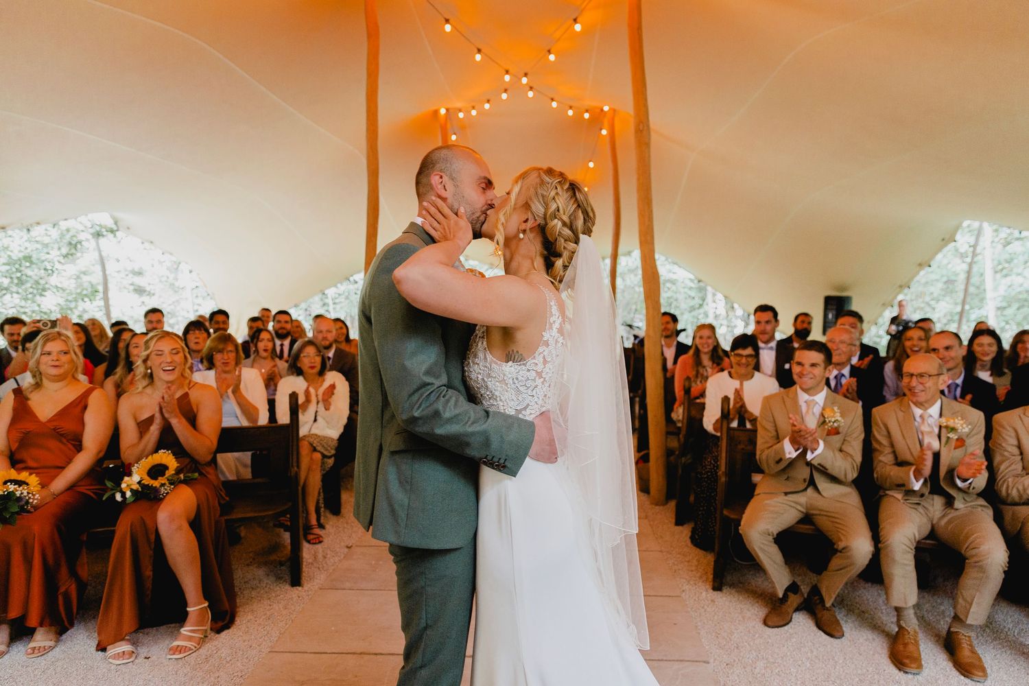 Bride and groom share first kiss at altar inside elegant tent ceremony with guests applauding.
