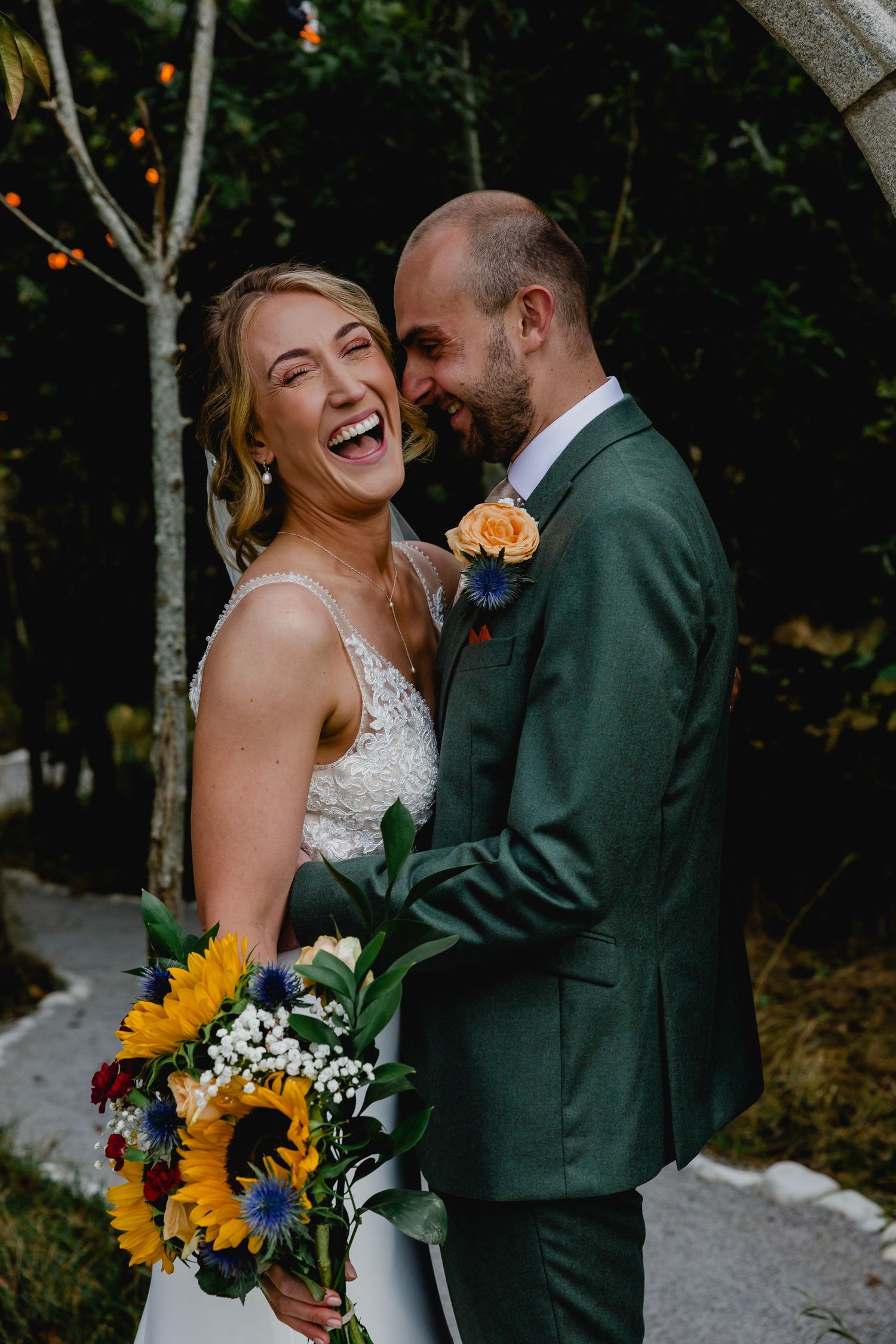 Joyful bride laughing with groom in green suit, holding vibrant sunflower bouquet during outdoor wedding ceremony.