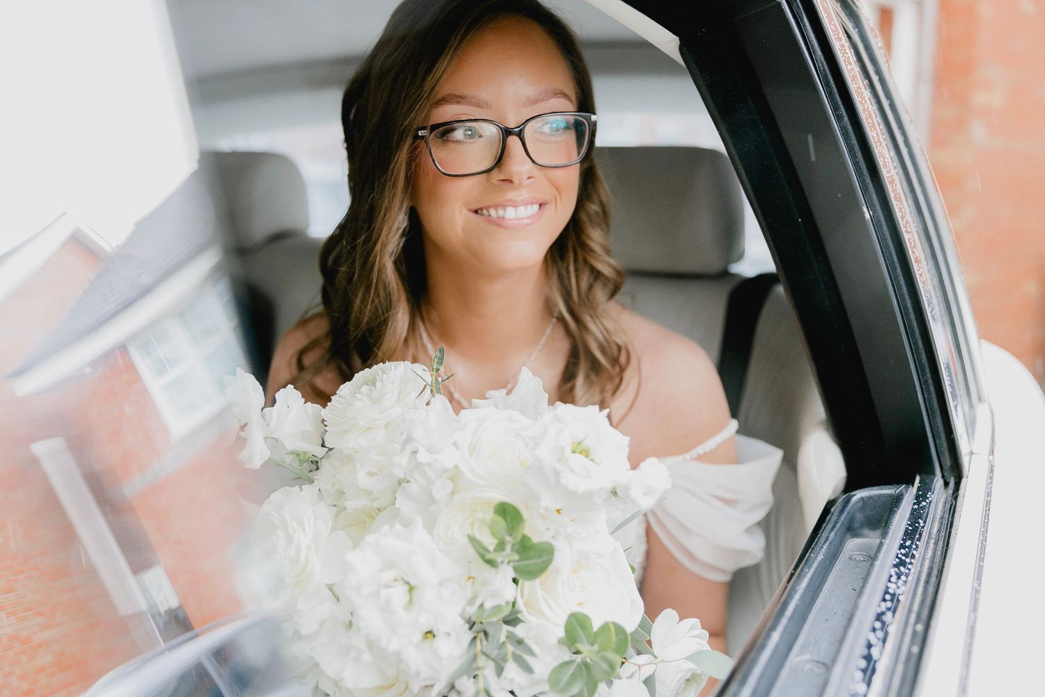 Smiling bride wearing glasses holding white floral bouquet while sitting in wedding car.