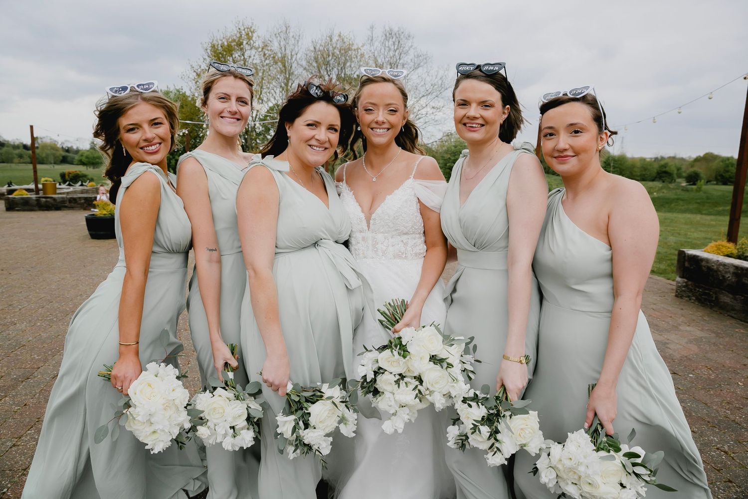 Bride with five bridesmaids in sage green dresses holding white bouquets at outdoor wedding.