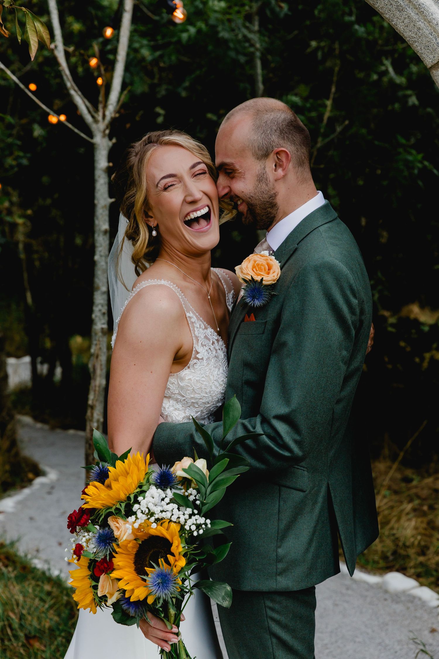 Laughing bride holding sunflower bouquet with groom in green suit embracing outdoors at woodland wedding.