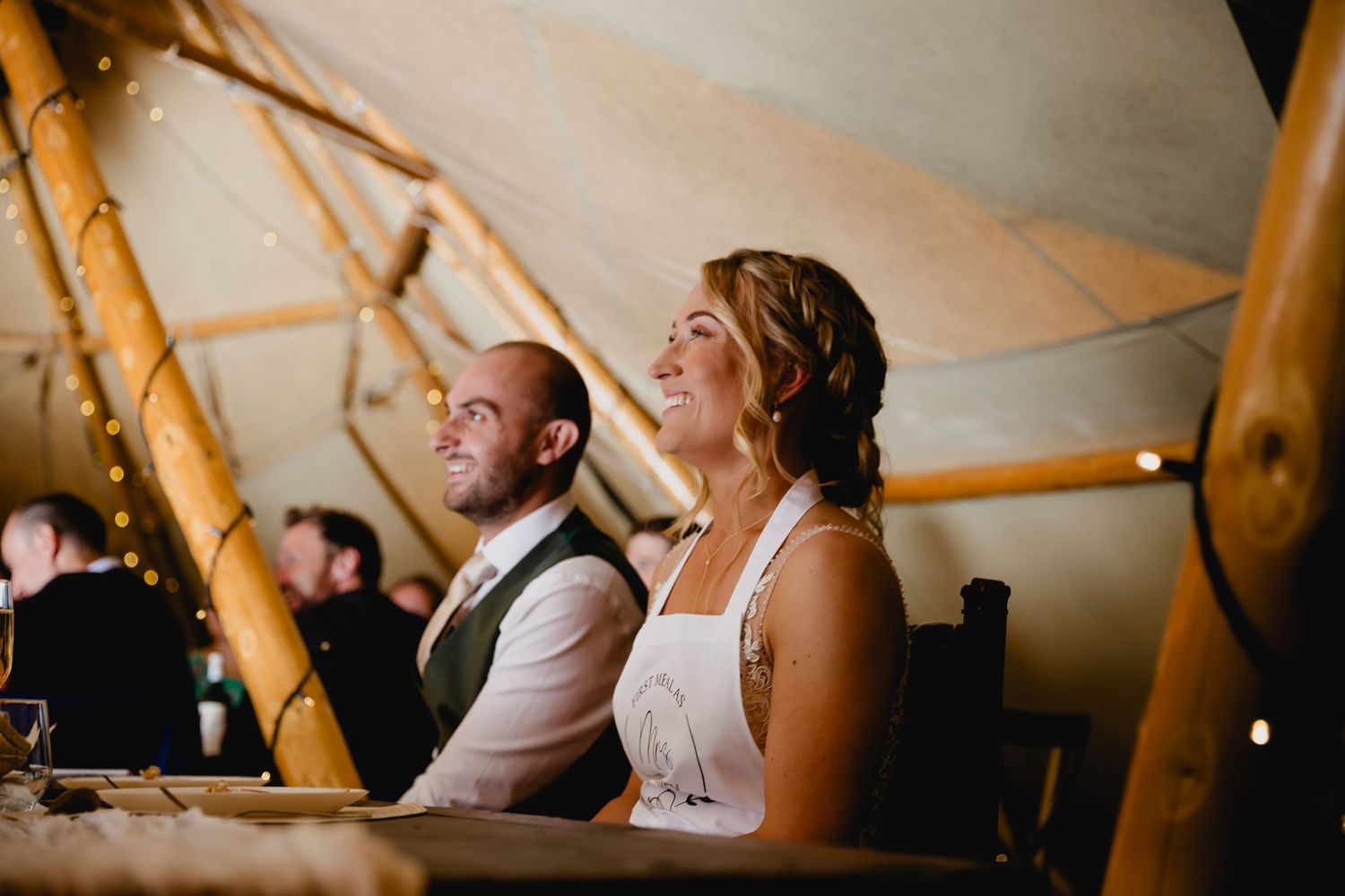 Bride and groom seated at head table inside warmly lit tipi reception venue.