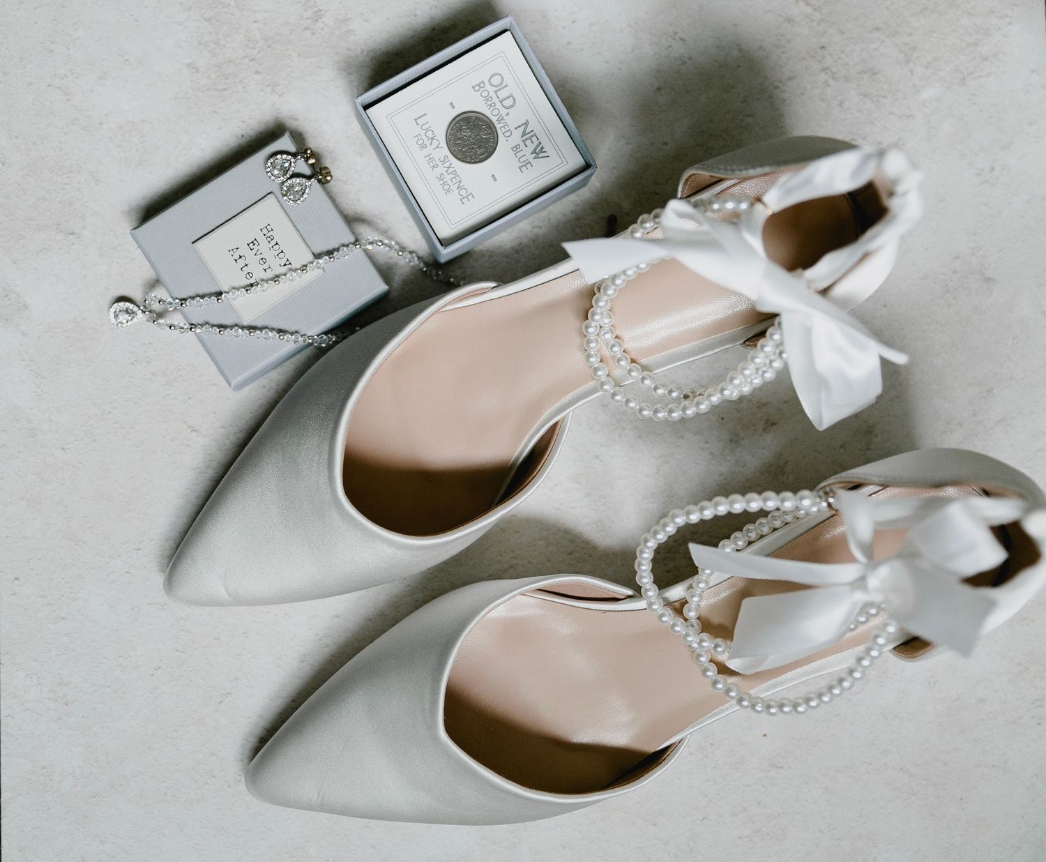 White satin bridal heels with pearl ankle straps styled with perfume and jewelry flatlay.