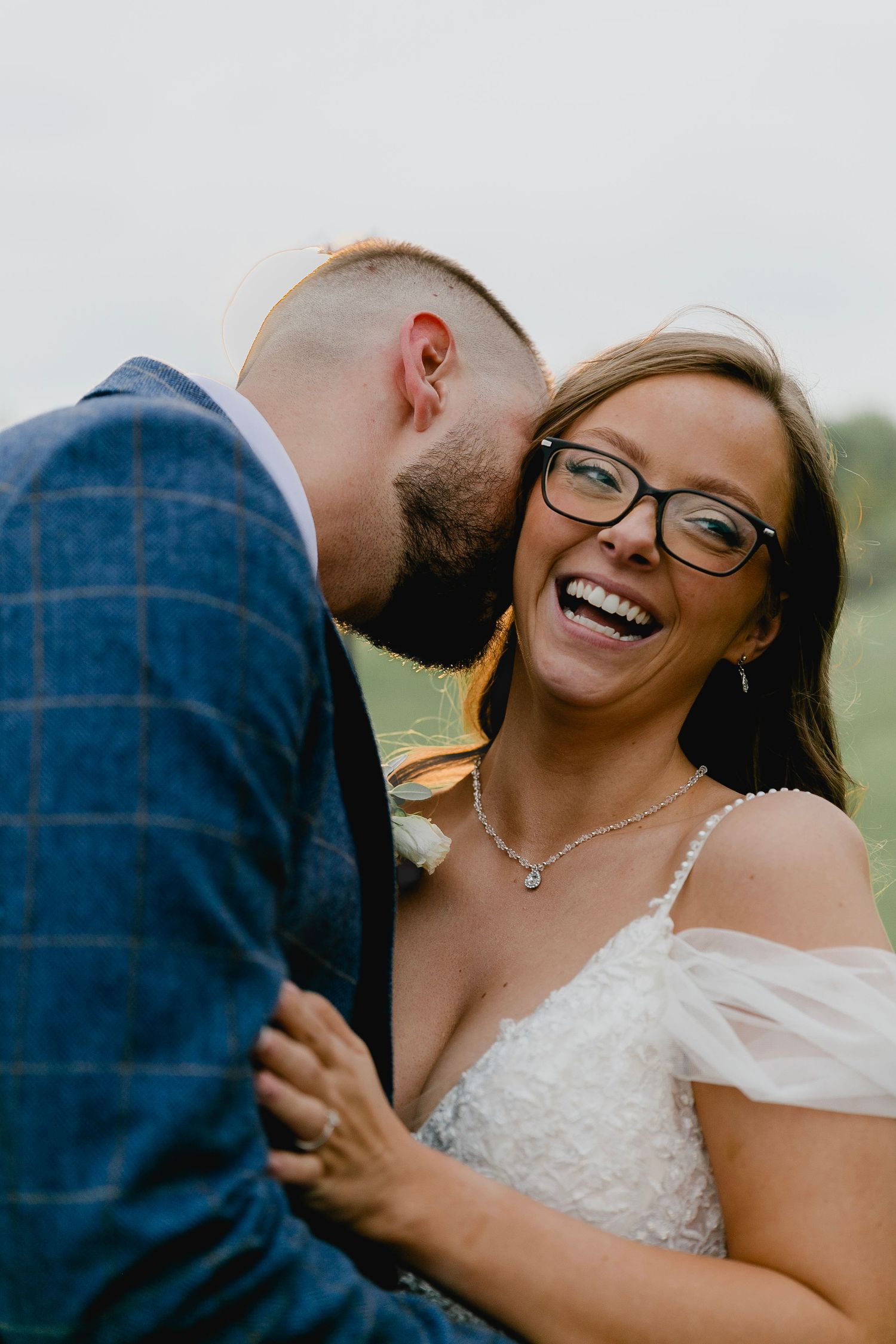 Bride in white lace gown laughing as groom whispers in her ear