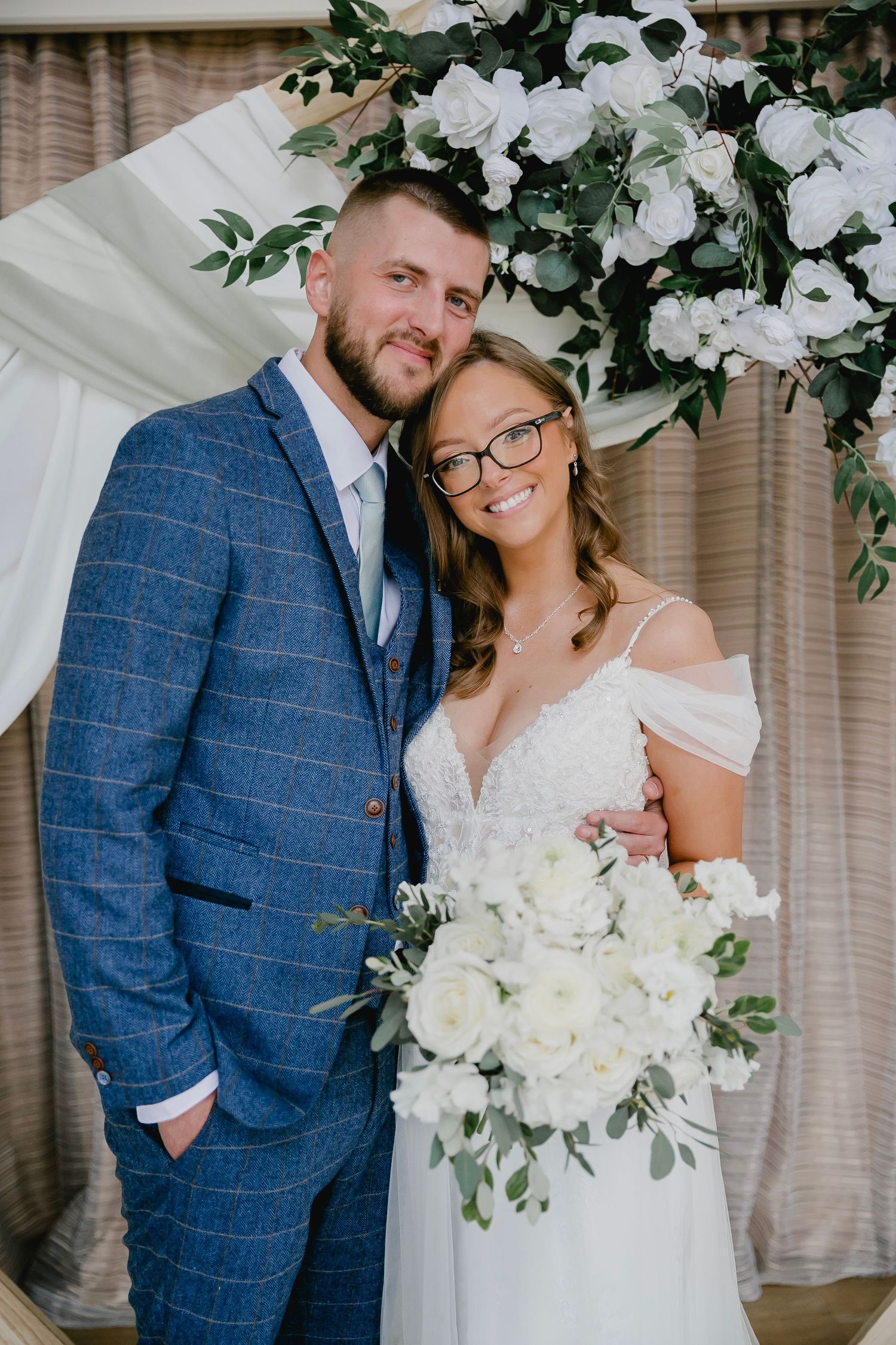 Smiling bride and groom posing before white floral arch, bride holding white bouquet.