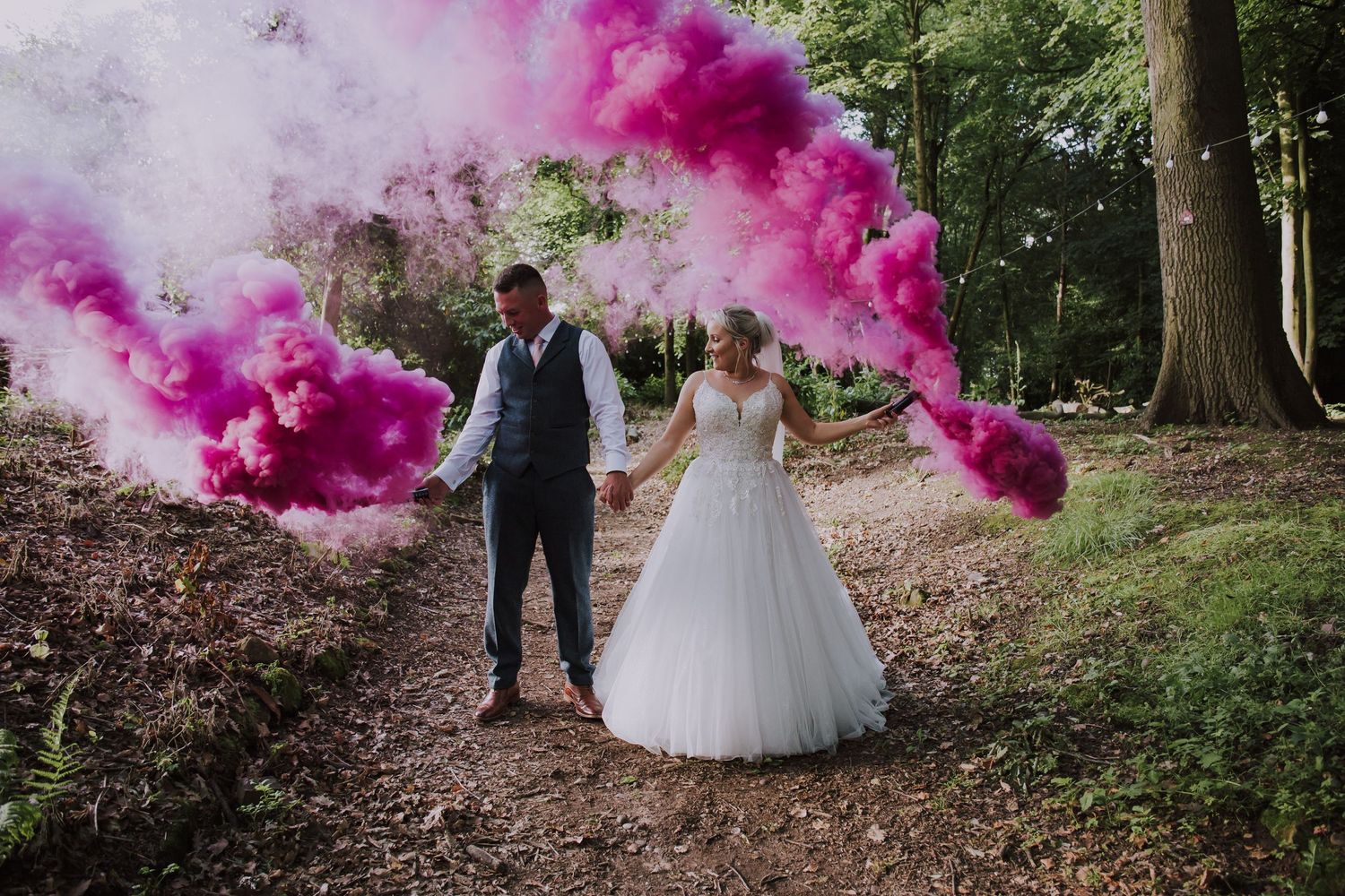 Bride and groom walking in forest holding pink smoke bomb on wedding day.
