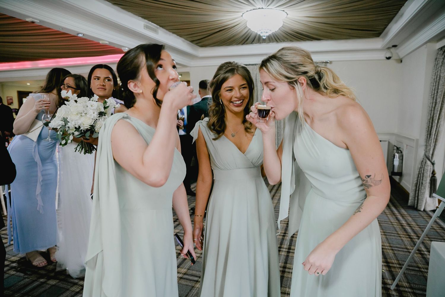 Three bridesmaids in sage green dresses laughing and drinking at wedding reception.