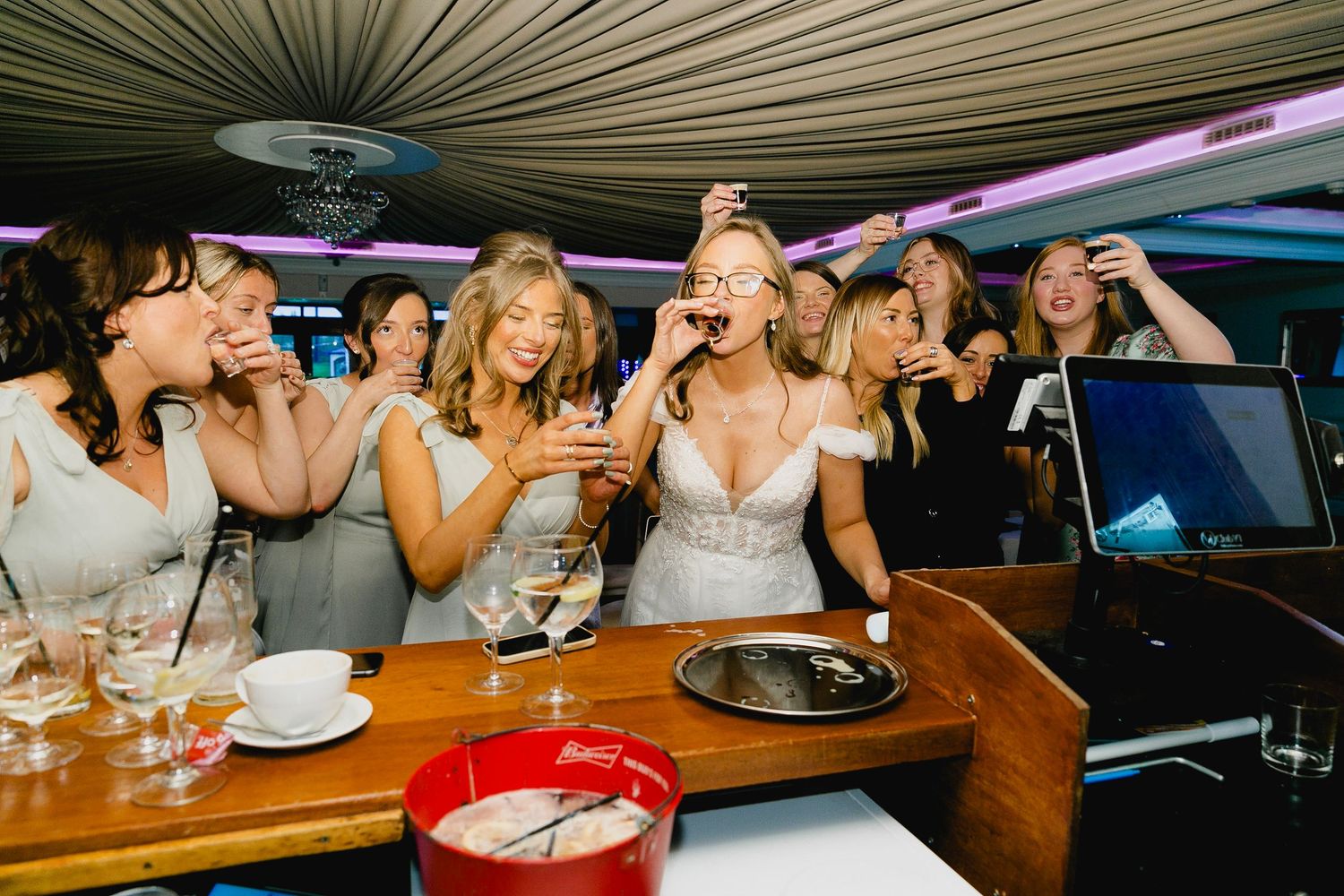 Bride and bridesmaids in sage dresses laughing and celebrating at wedding reception bar.