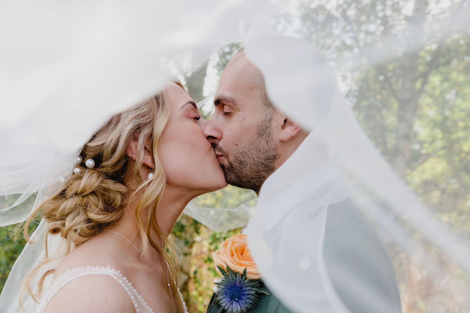 Bride and groom sharing a kiss under flowing white veil with orange and blue wedding flowers.