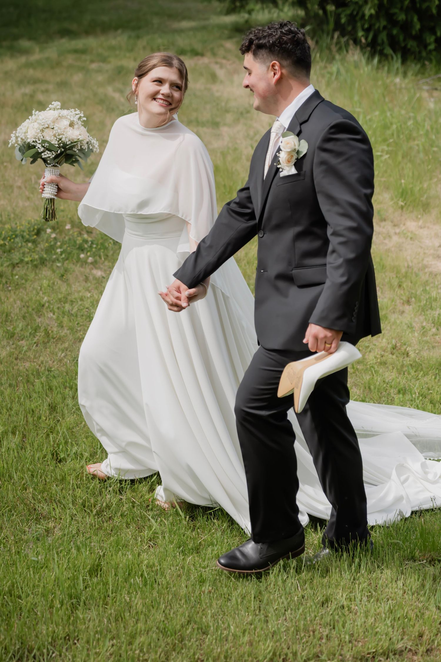 Bride in white cape gown holds bouquet while groom in dark suit holds her hand, walking on green grass.