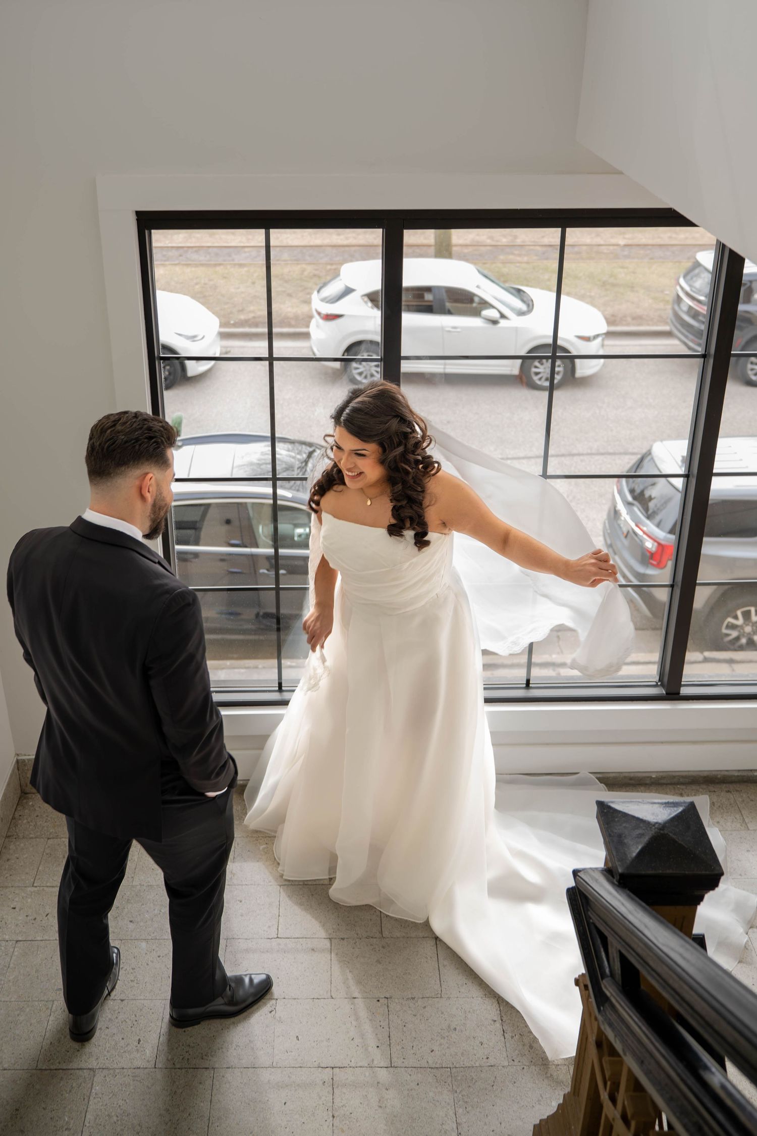 Bride in white gown and groom in black suit share a moment by large windows indoors.