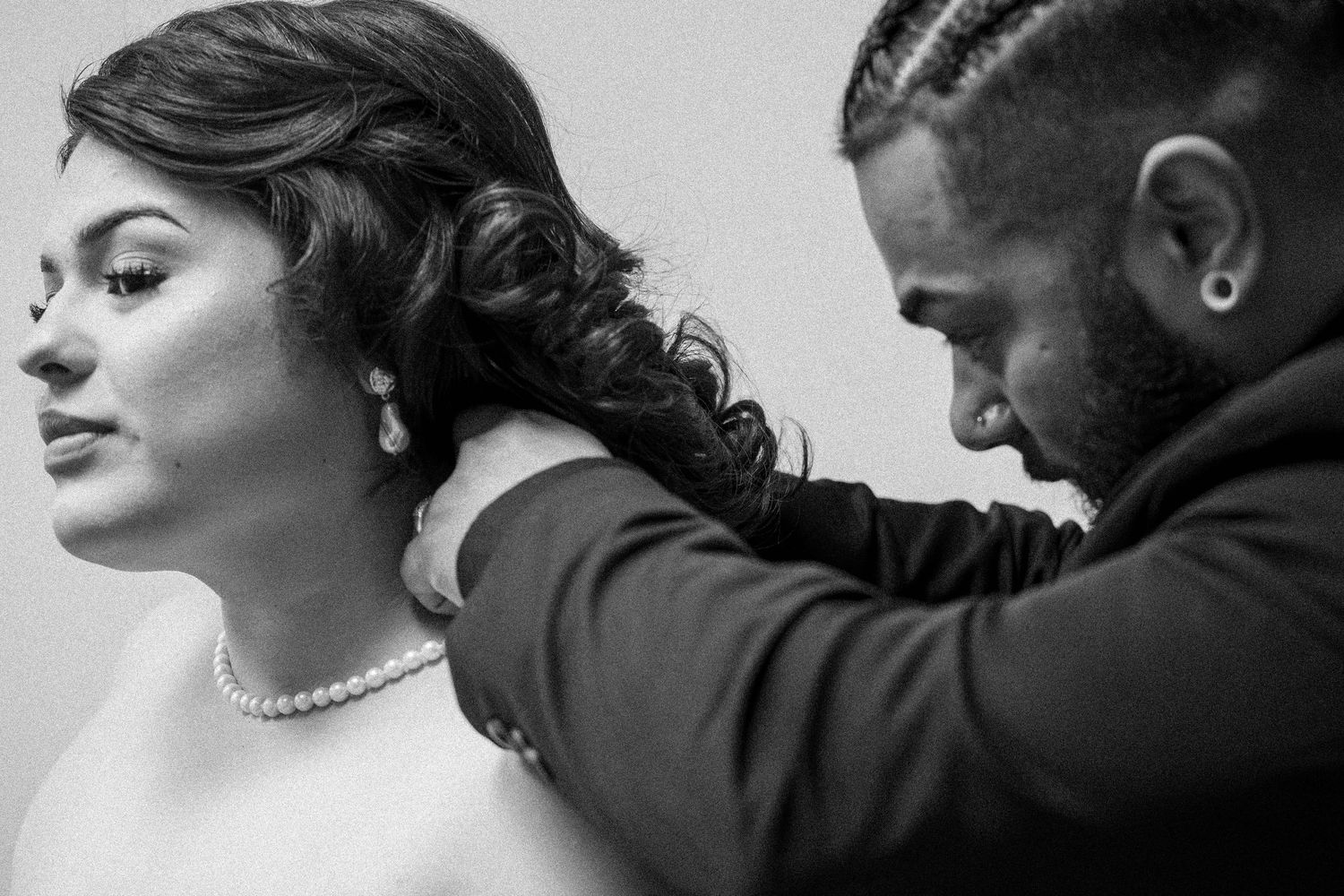 Groom with braided hair fastens pearl necklace on bride in black and white wedding portrait.