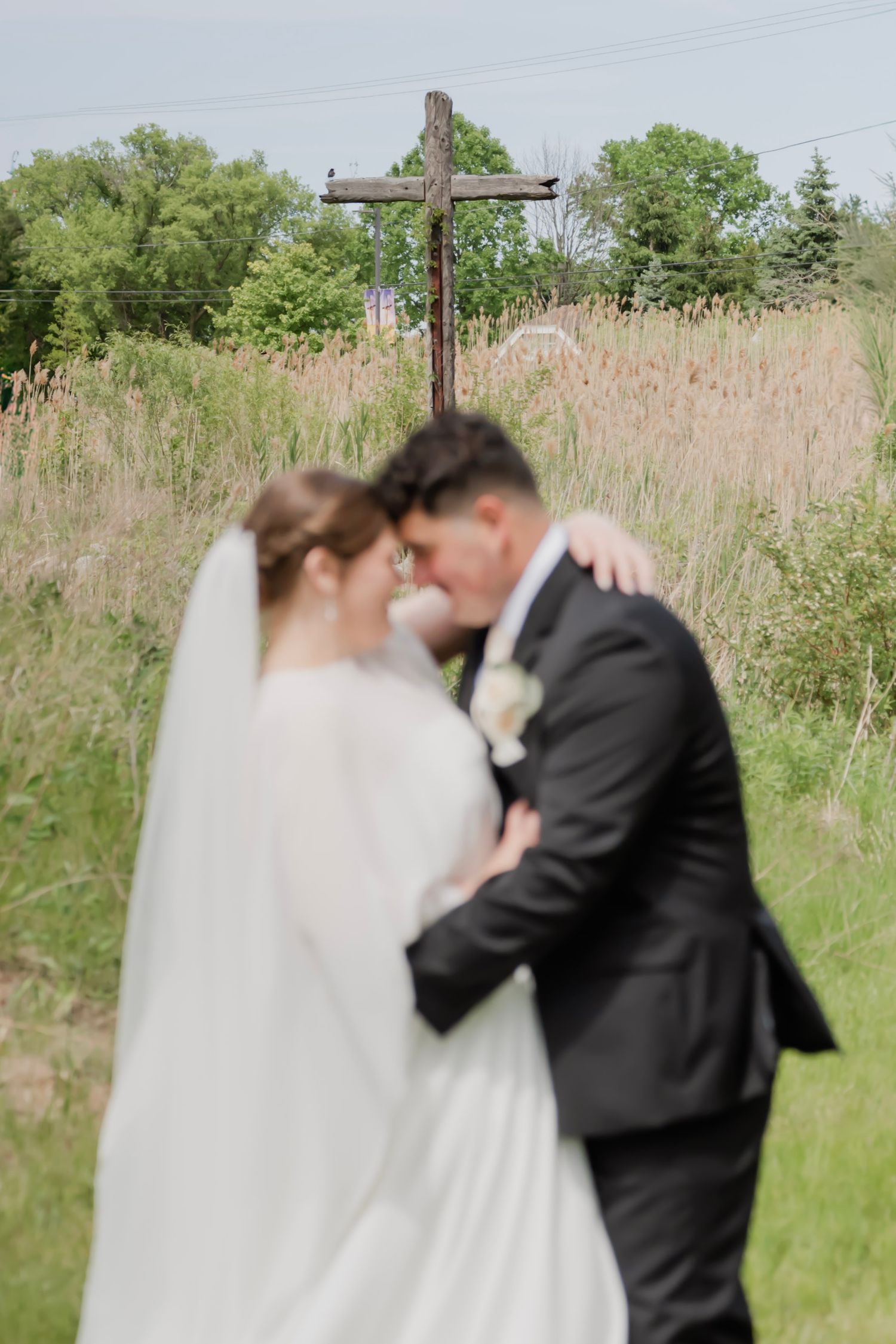 Bride and groom embrace in front of a wooden cross in a lush green field on their wedding day.