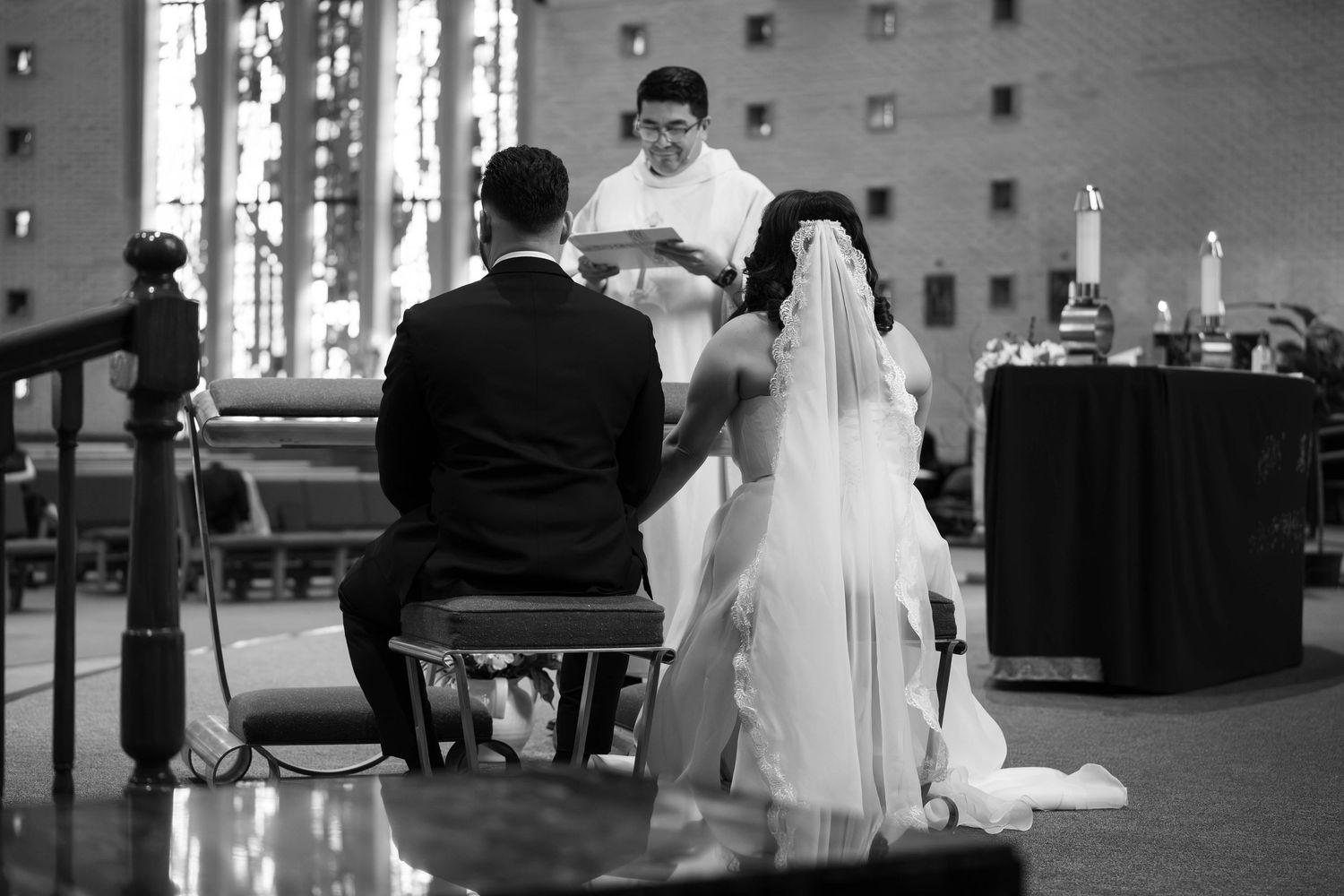 Black and white wedding ceremony photo of bride and groom kneeling before officiant at church altar.
