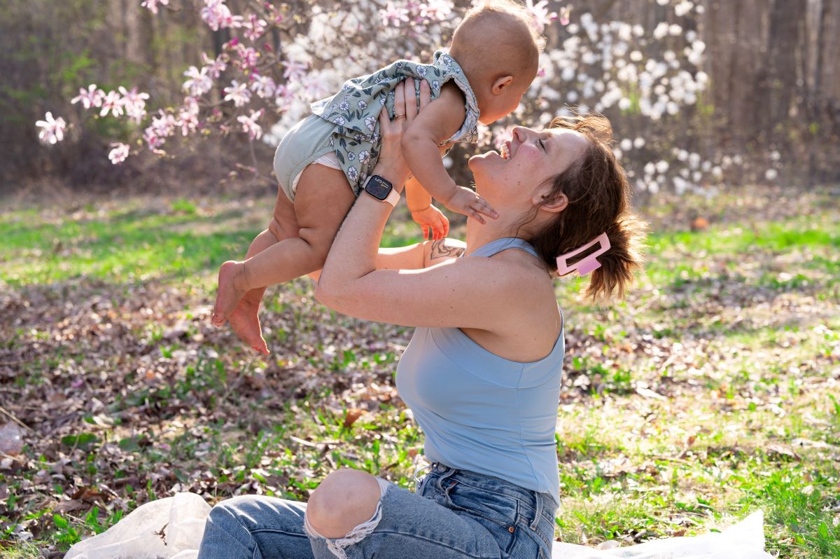 Joyful mother lifts smiling baby in the air outdoors near blooming spring trees.