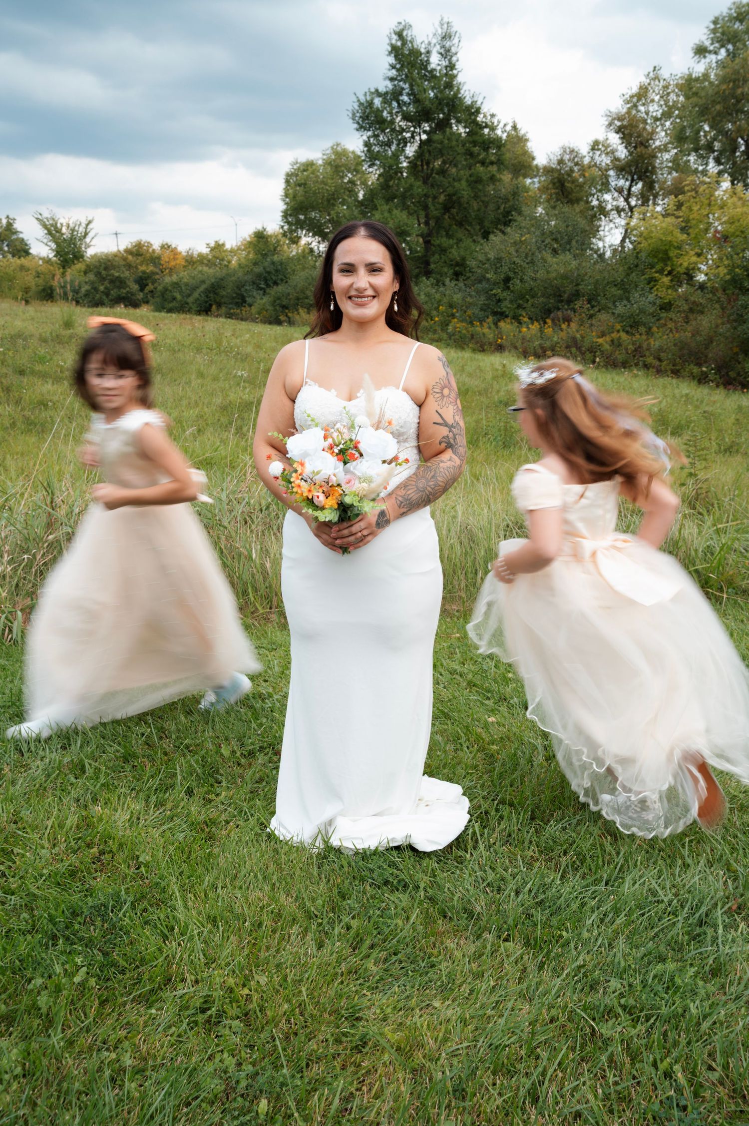 Bride in white gown holds bouquet while flower girls in blush dresses twirl around her in a green meadow.