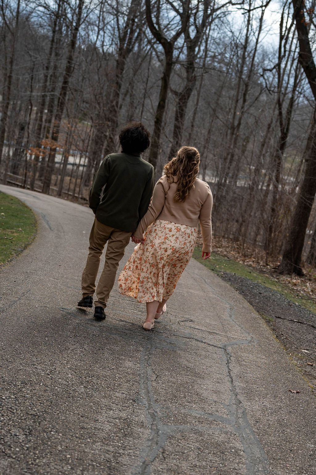 A couple holds hands while walking away on a winding path surrounded by bare trees in a park.