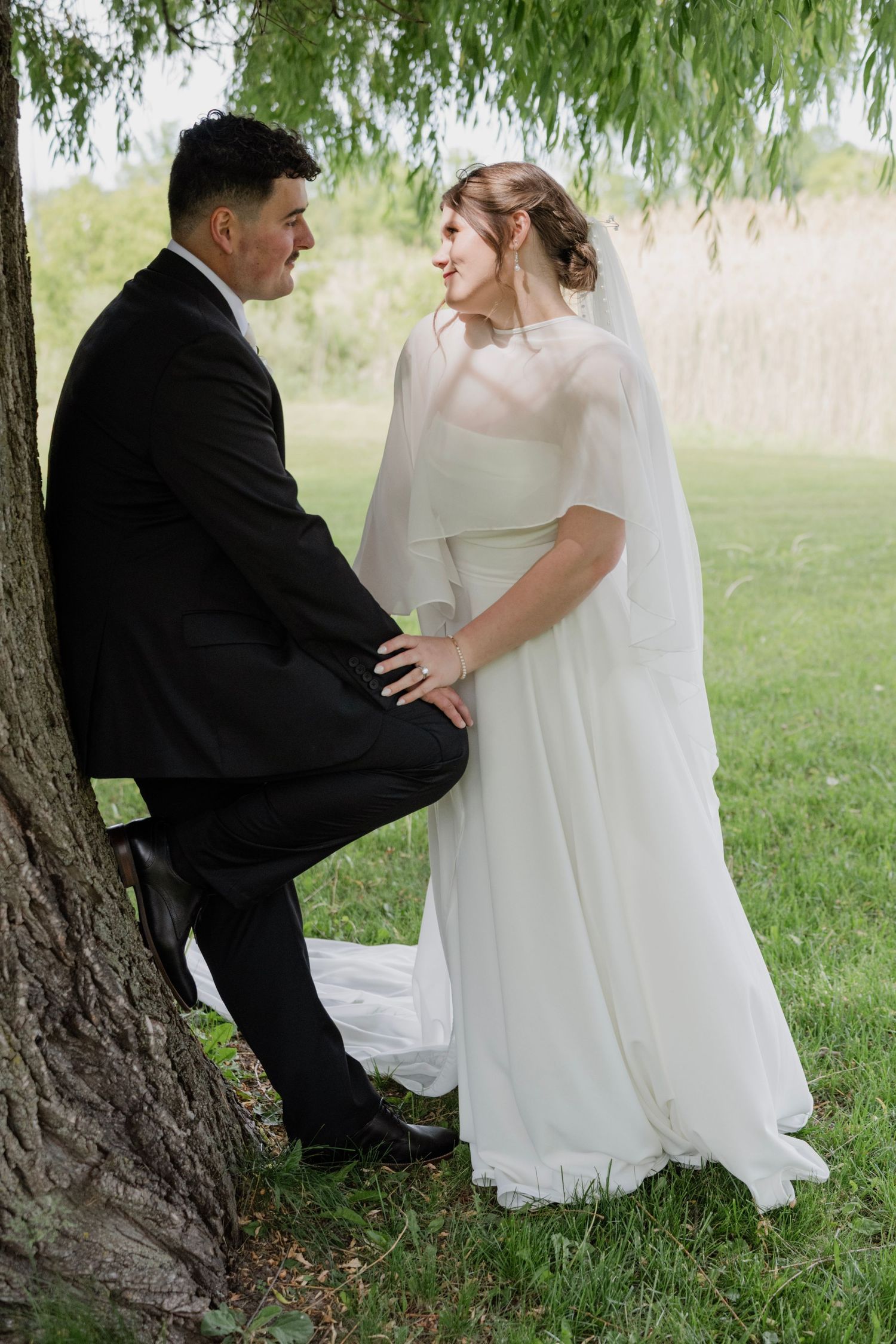 Bride in white cape gown and groom in black suit share a romantic moment leaning against a tree outdoors.