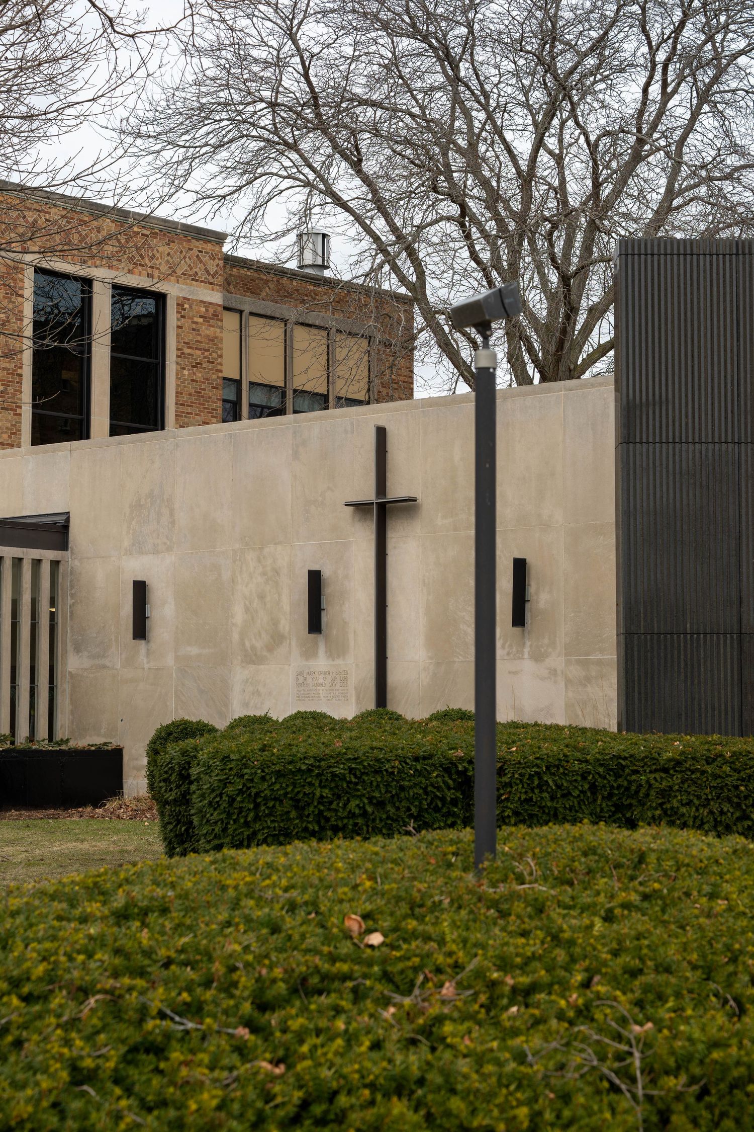 Modern church exterior with concrete wall featuring a cross, trimmed hedges, and bare trees against cloudy sky.