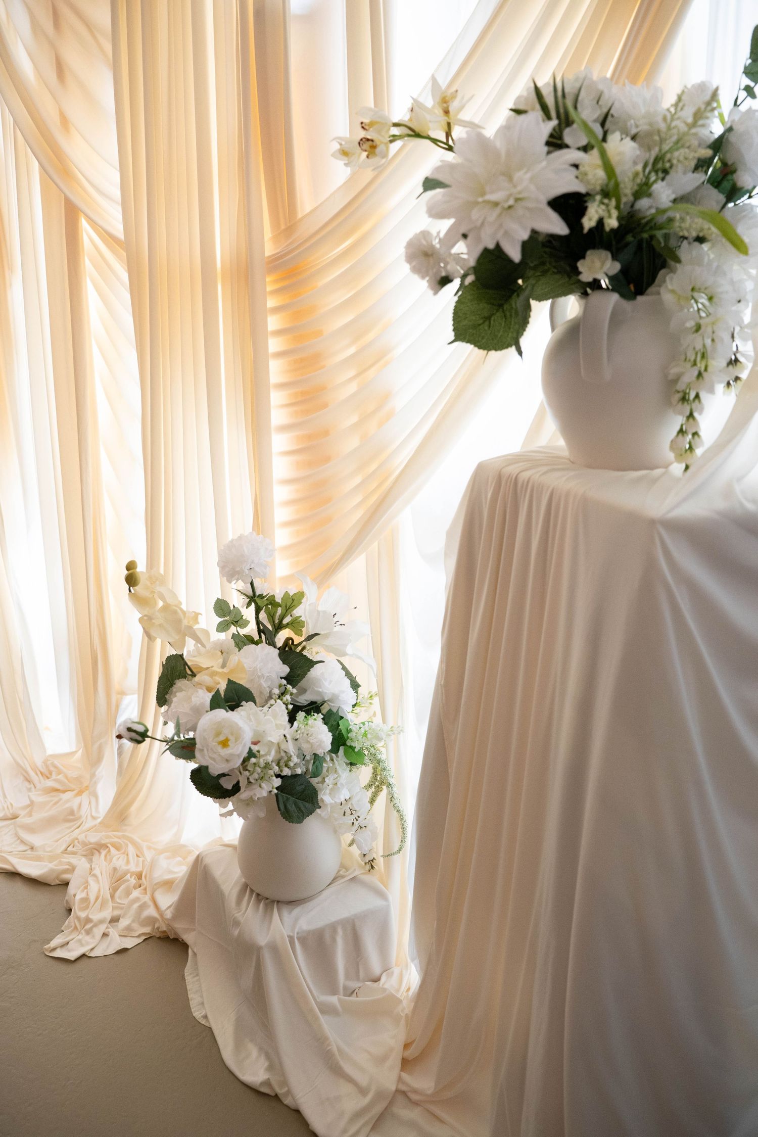 White wedding floral arrangements in ceramic vases displayed against draped cream fabric backdrop.