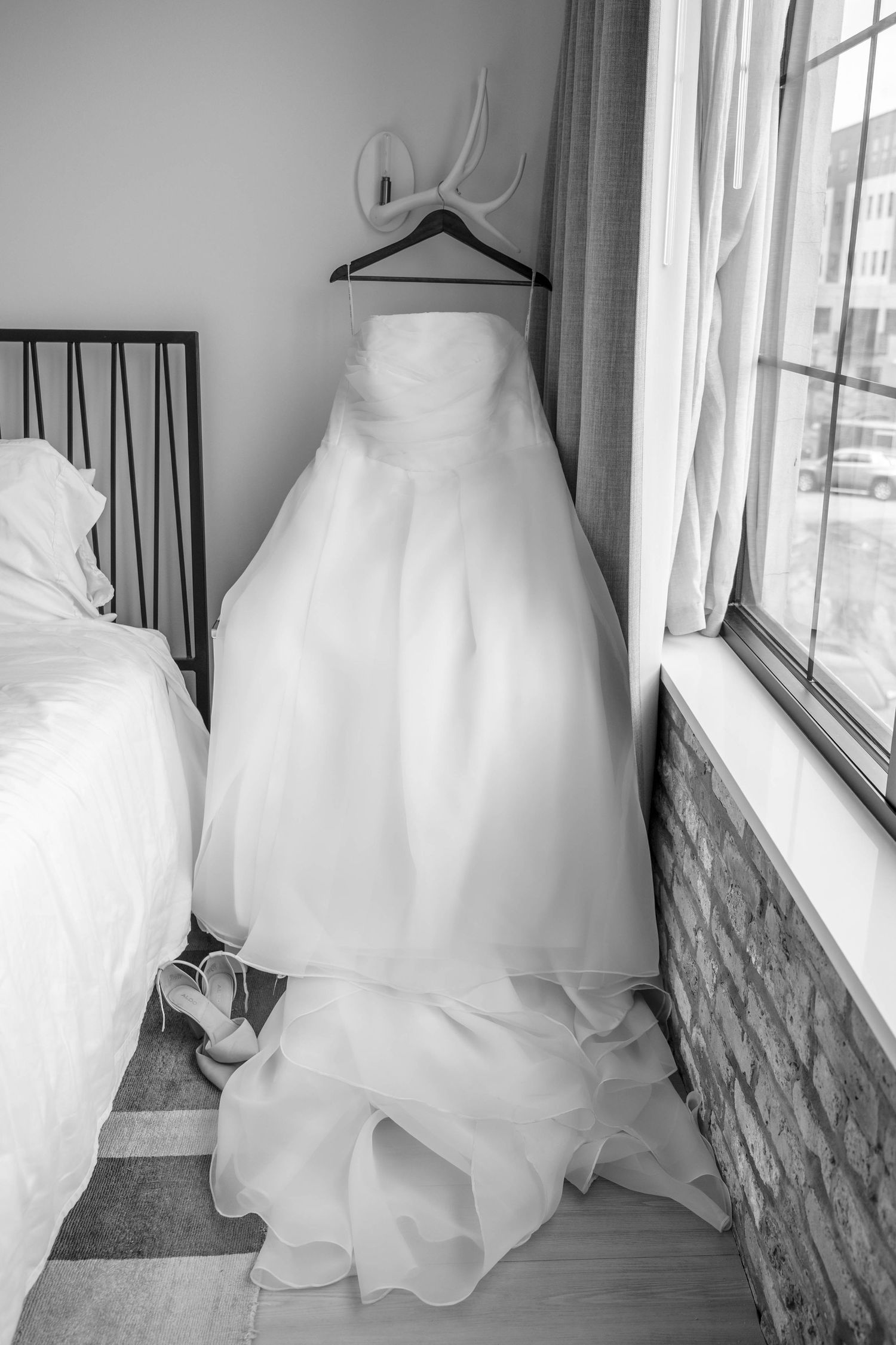 Black and white photo of elegant strapless wedding gown hanging near window with bridal heels below.