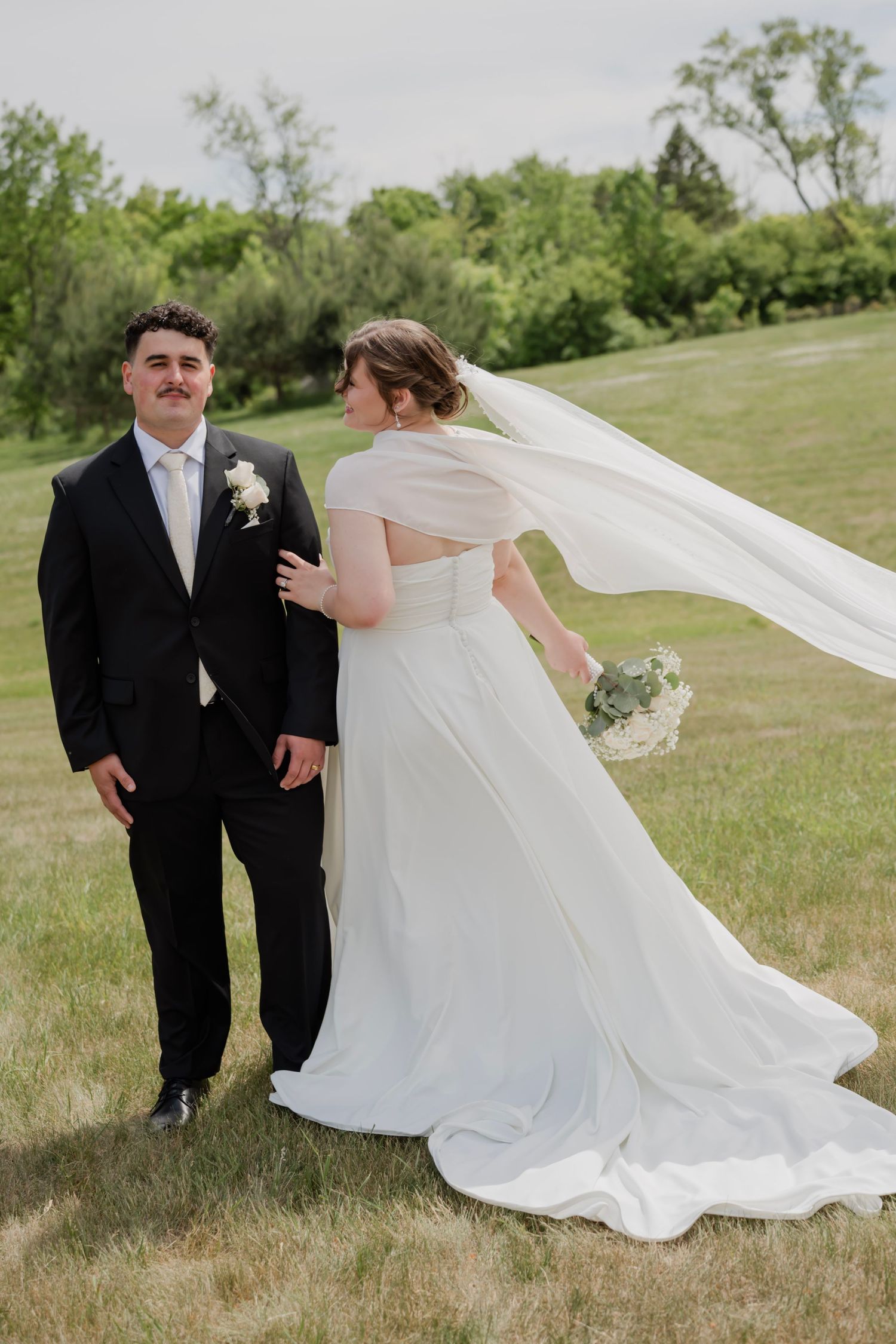 A bride in a flowing white gown with veil blowing in wind stands with groom in black suit on green grass.