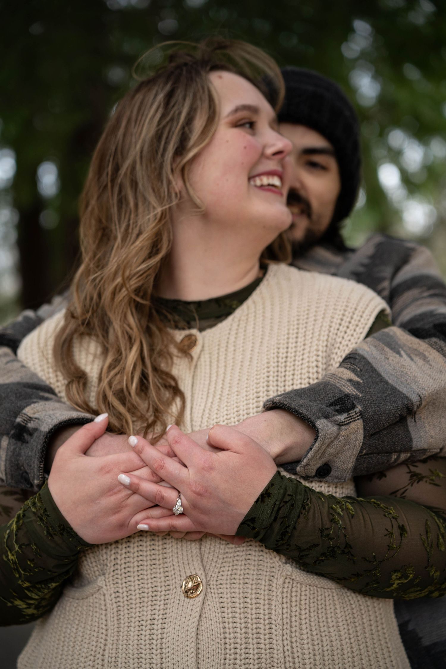 Smiling woman in cream sweater vest embraced from behind by man in flannel, showing engagement ring outdoors.