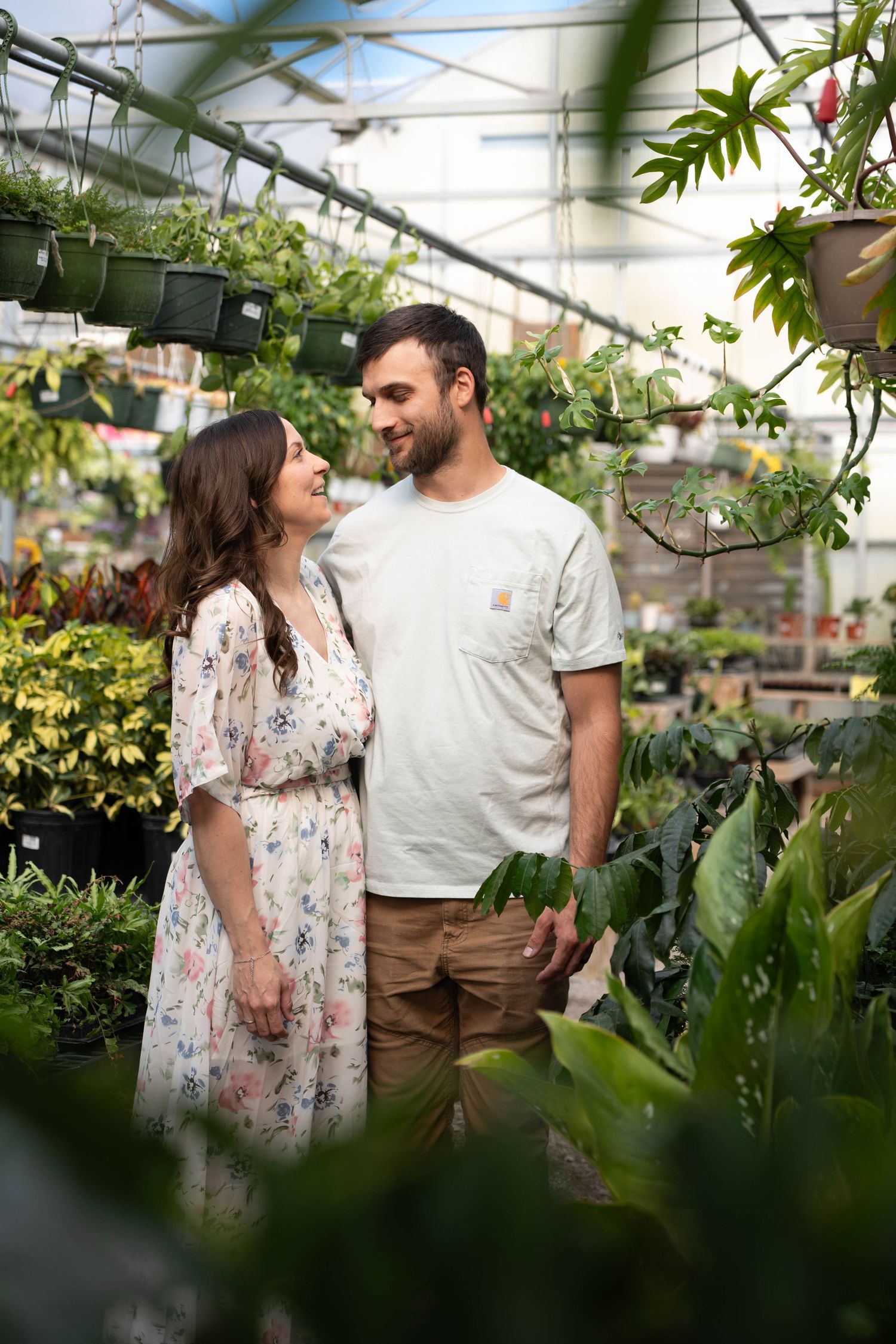 Happy couple smiling at each other surrounded by lush green plants in a greenhouse.