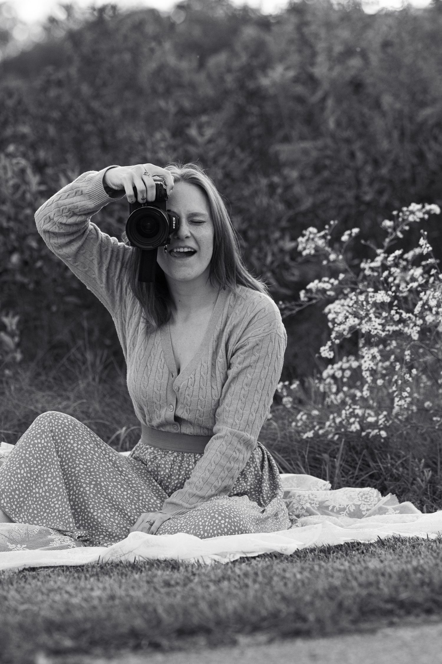 Black and white portrait of young woman sitting on blanket outdoors holding a camera.