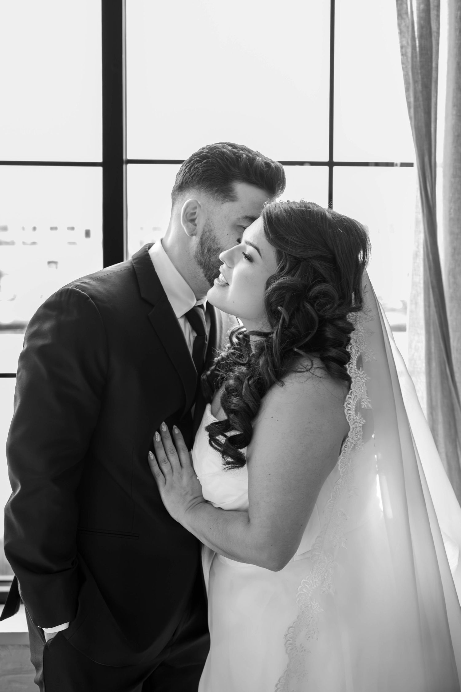 Black and white photo of bride and groom sharing an intimate moment near large windows on wedding day.