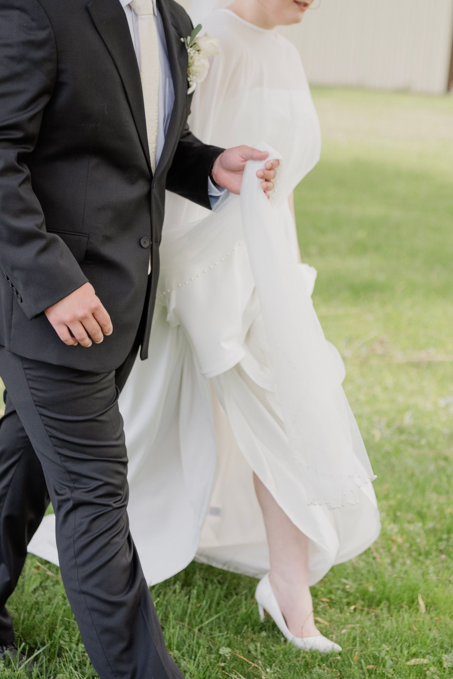 Bride in flowing white gown walks arm-in-arm with groom in dark suit on green grass at outdoor wedding.