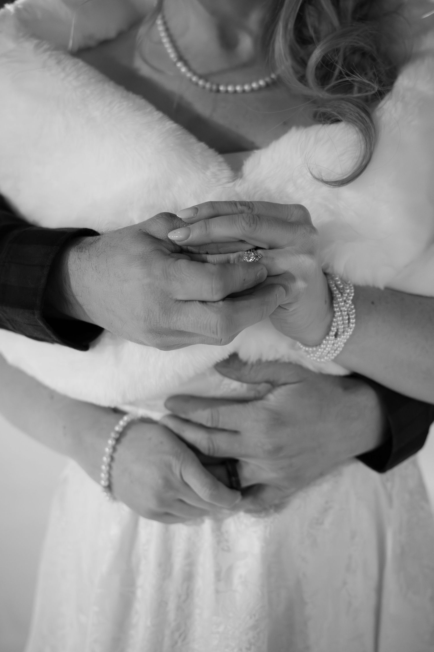 Close-up black and white photo of bride and groom holding hands showing diamond ring and pearl bracelets.