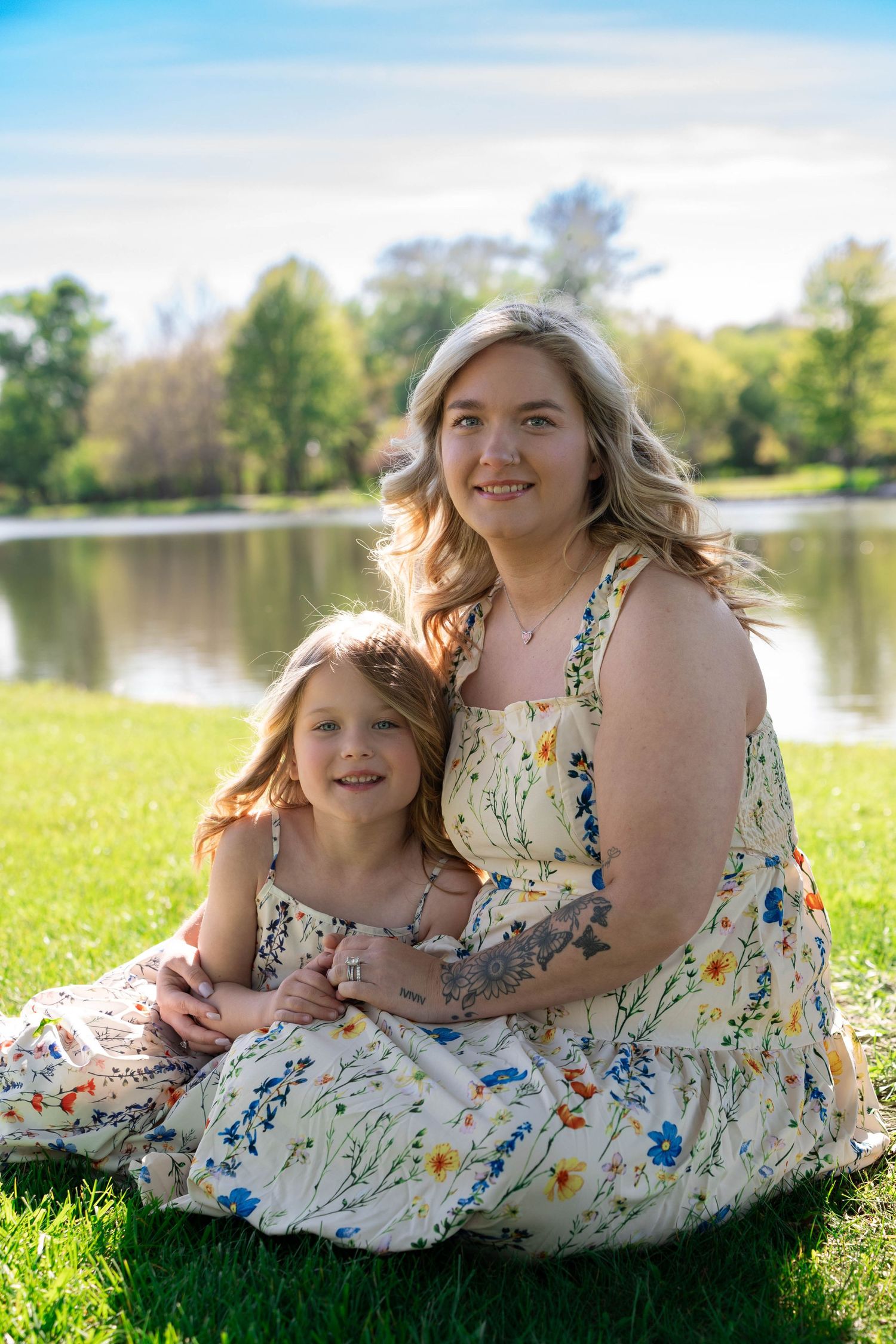 Mother and daughter in matching floral sundresses sit together on grass beside a peaceful lake.