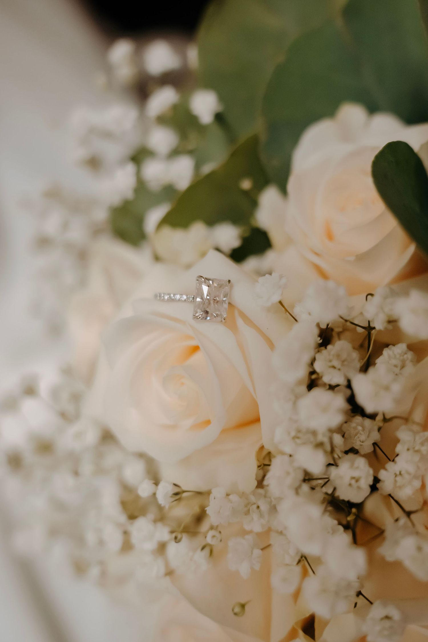 Diamond engagement ring resting on a bridal bouquet of white roses and baby's breath with green eucalyptus.