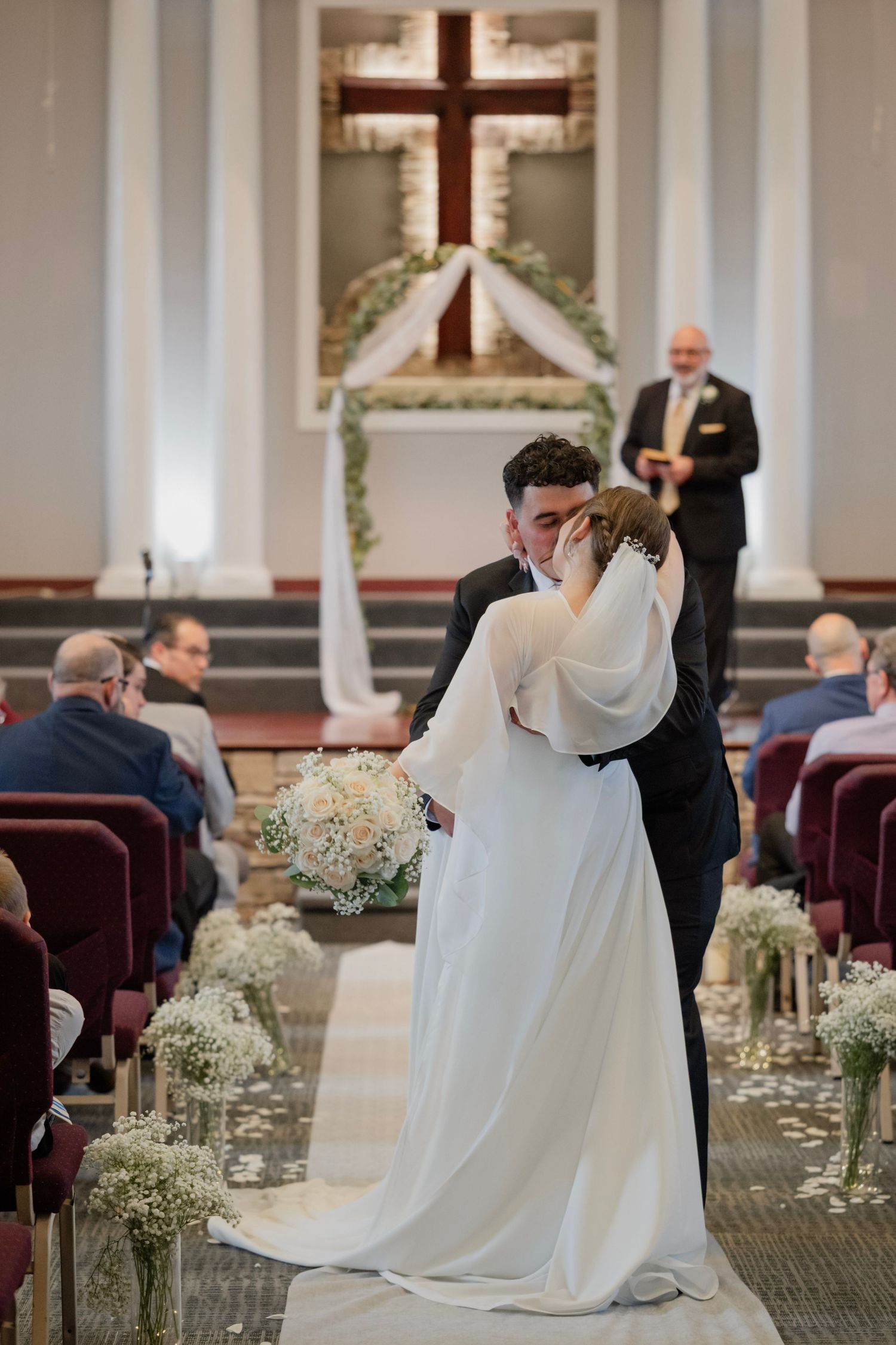 Bride and groom share first kiss at church wedding altar, bride holding white floral bouquet with guests seated in burgundy pews.