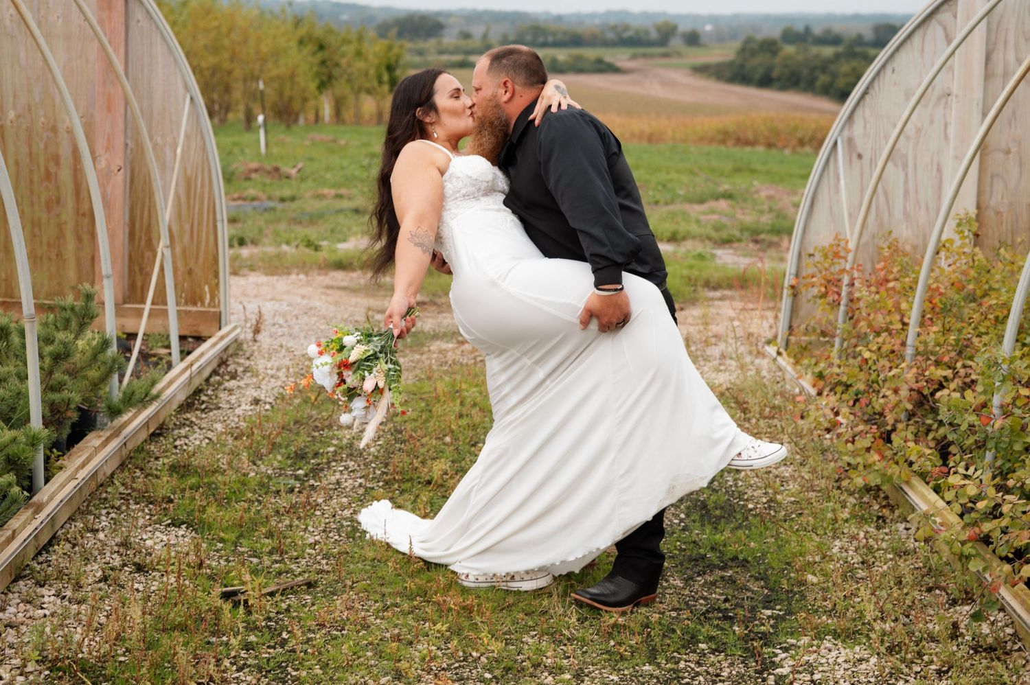 Groom dips bride for romantic kiss near greenhouse at rustic outdoor wedding.