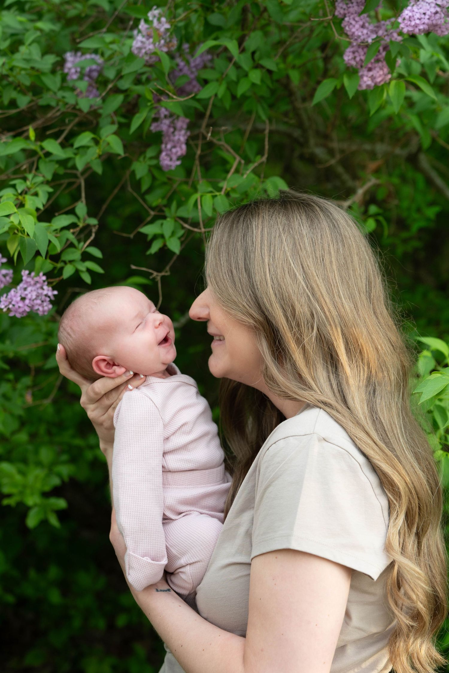 Blonde mother smiles lovingly at her newborn baby held in arms near blooming lilac bushes.